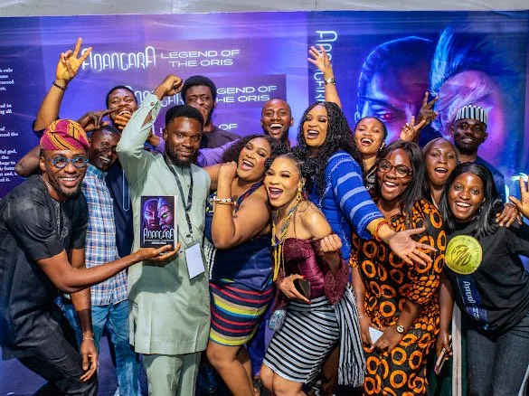 Group of people, celebrating and smiling at an event, with a movie poster for 'Ajancara: Legend of the Oris' in the background.