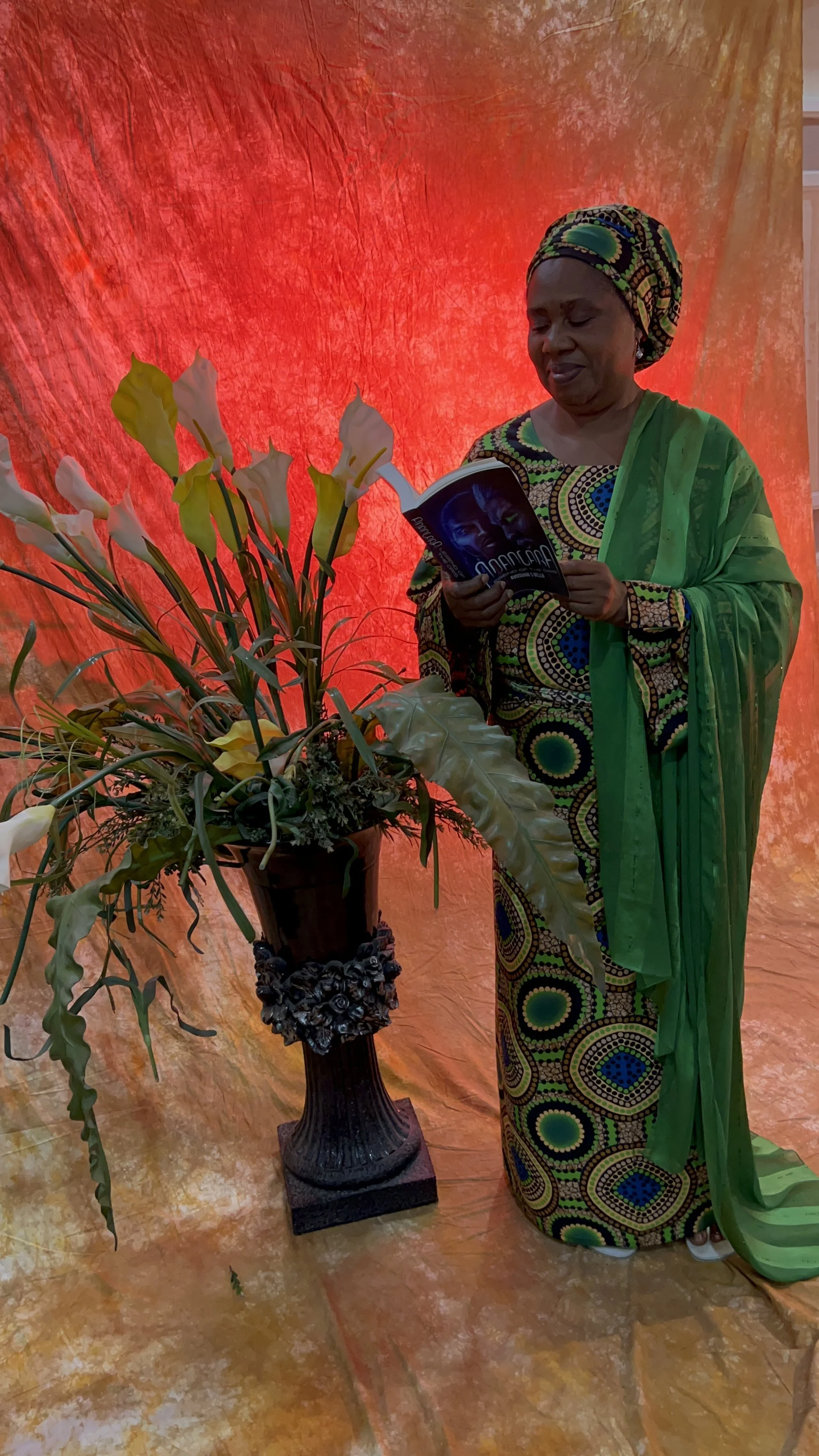 A woman wearing a colorful patterned African dress and head wrap is standing next to a large flower arrangement in a decorative dark vase, reading a book against a vibrant orange and red background.