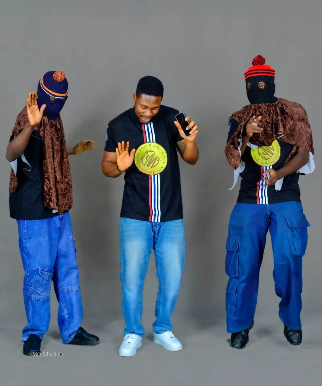 Three men dancing and having fun against a gray background. One in the middle is smiling and looking at his phone, while the two on the sides are wearing ski masks and costumes.