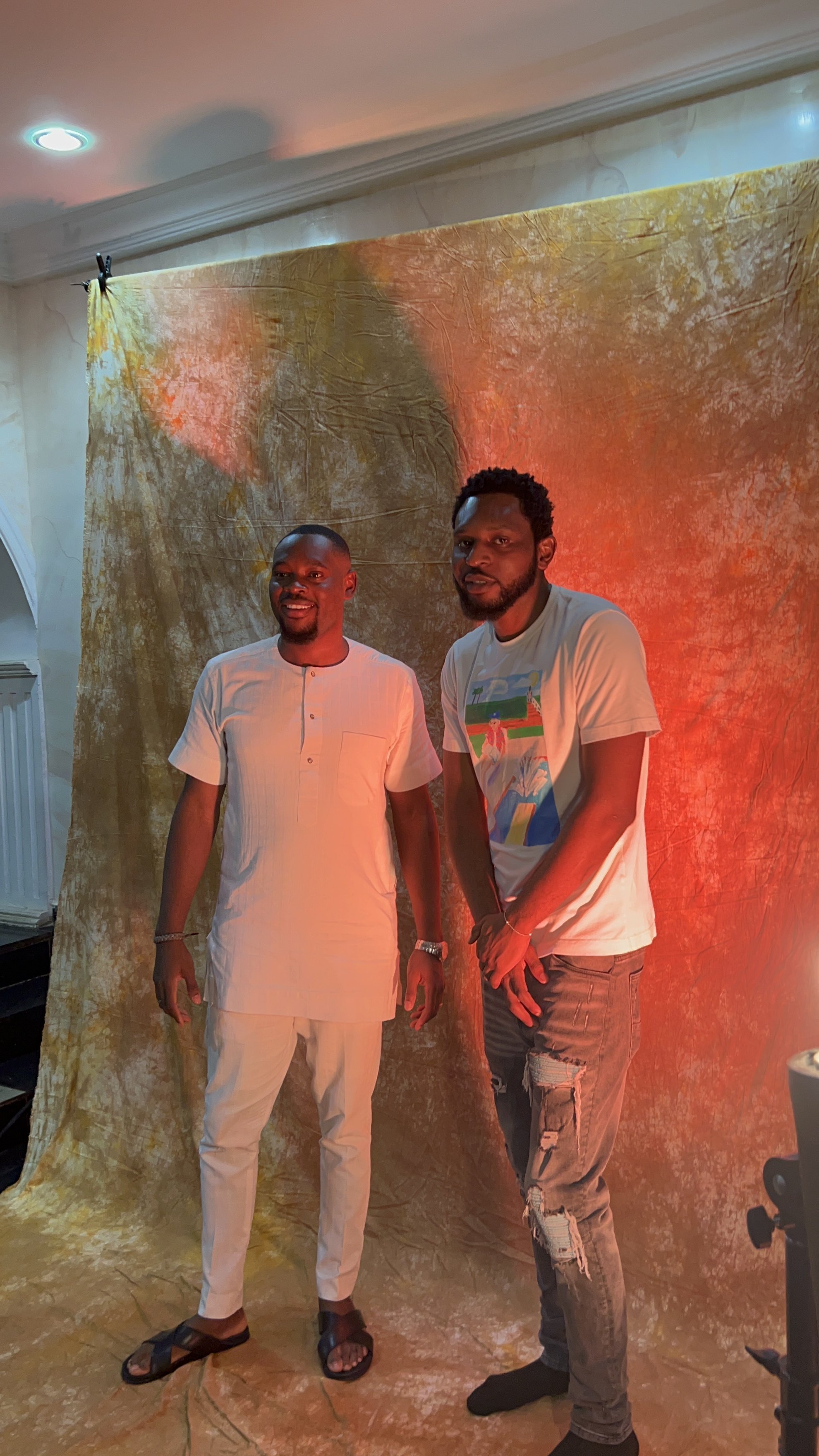 Two men standing in front of a colorful backdrop, one wearing a white traditional outfit and sandals, the other in a graphic t-shirt and ripped jeans, with warm lighting in the background.