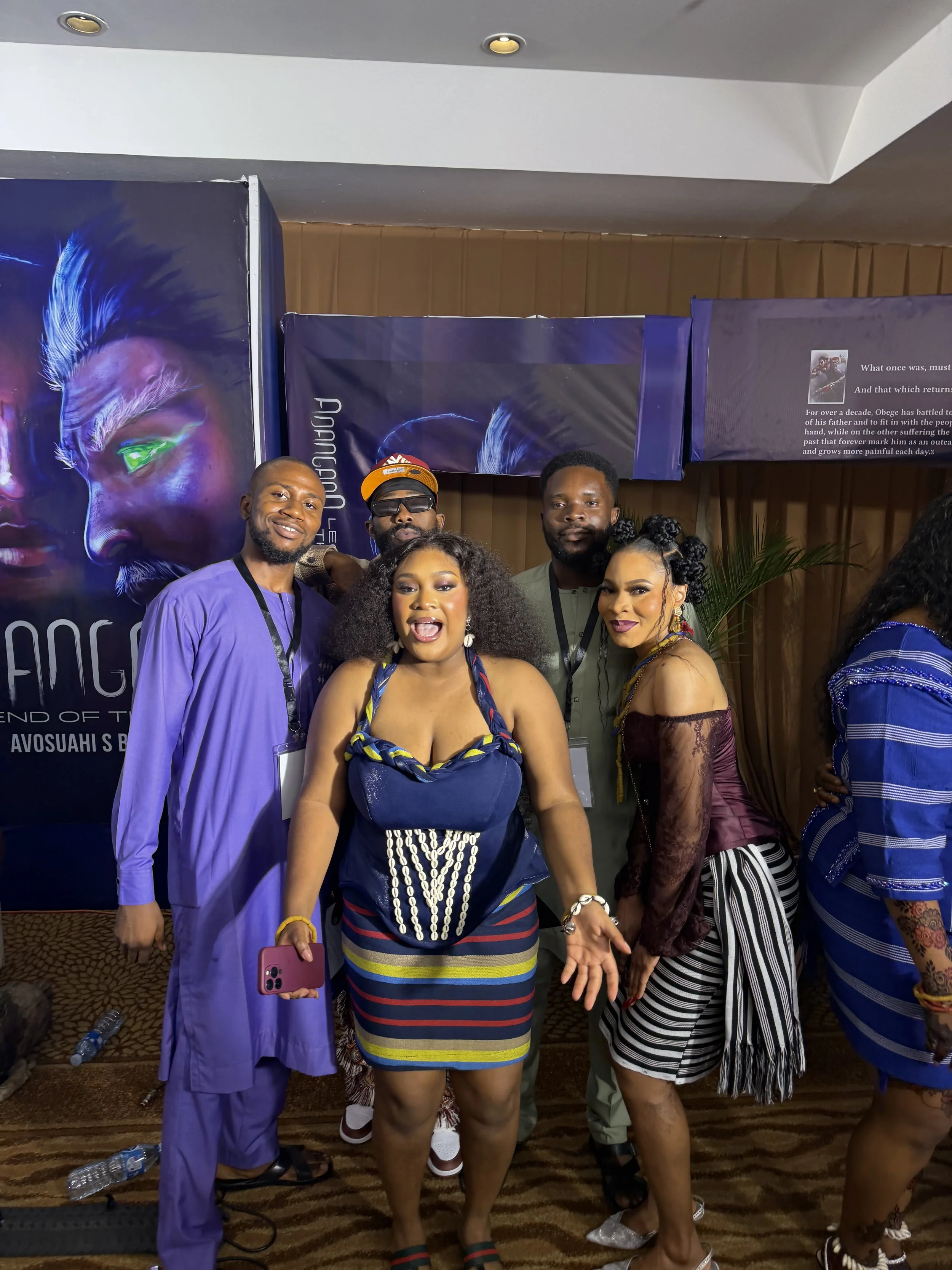 A group of five people posing together indoors at a promotional event, with posters and banners in the background. The woman in the center is wearing a colorful dress and accessorized, holding a smartphone. The others are dressed in colorful clothing, smiling, and standing close together.