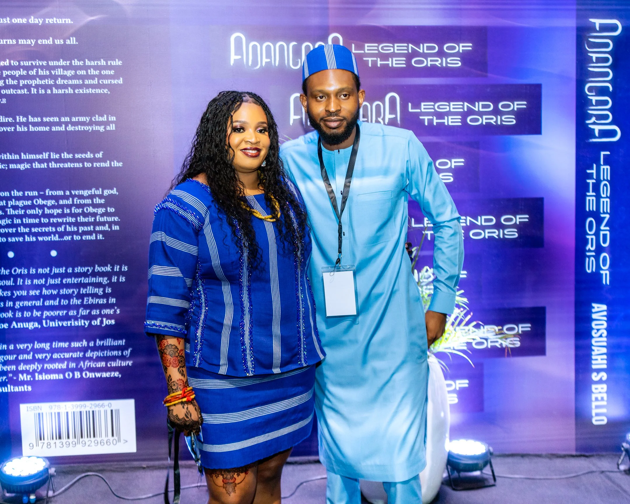 Two people standing in front of a purple book display background. The woman on the left wears a blue striped dress and has long curly black hair. The man on the right wears traditional light blue attire with a cap. Both are smiling and facing the camera.