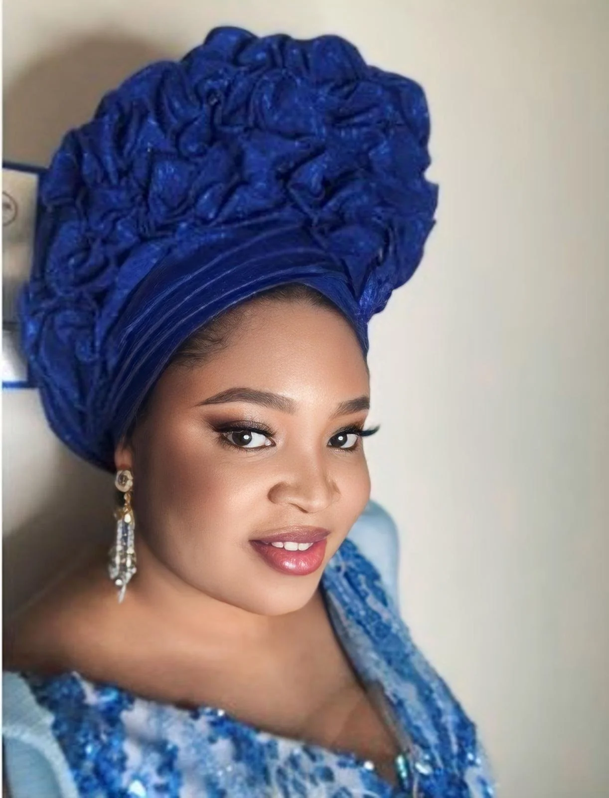 A woman dressed in traditional Nigerian attire with a large, elaborate blue gele headwrap, matching blue dress, and elegant earrings.