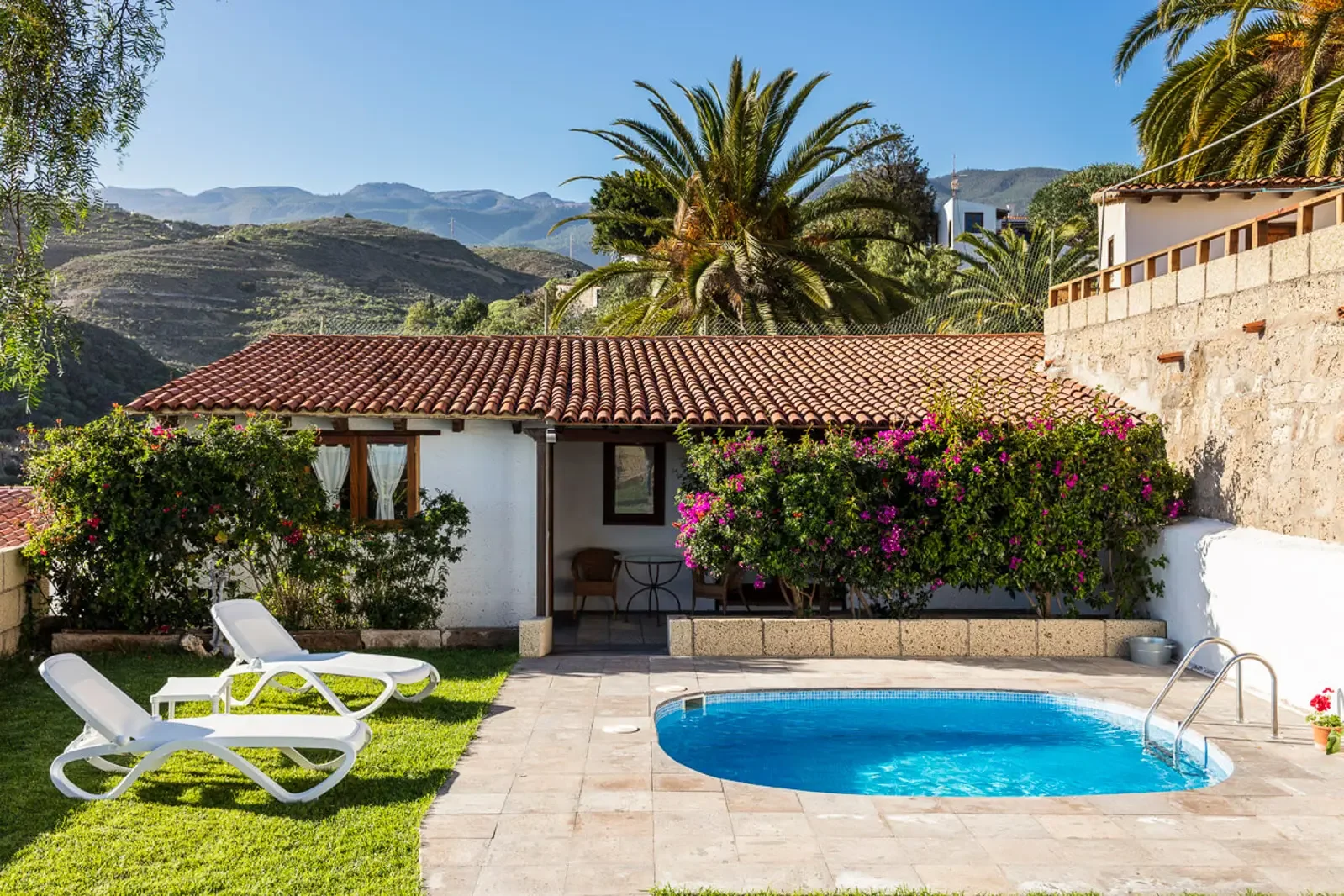eine kleine Villa mit einem roten Ziegeldach, ein Garten mit Pool, Liegestühlen und bunten Blumen, umgeben von Bergen und Palmen