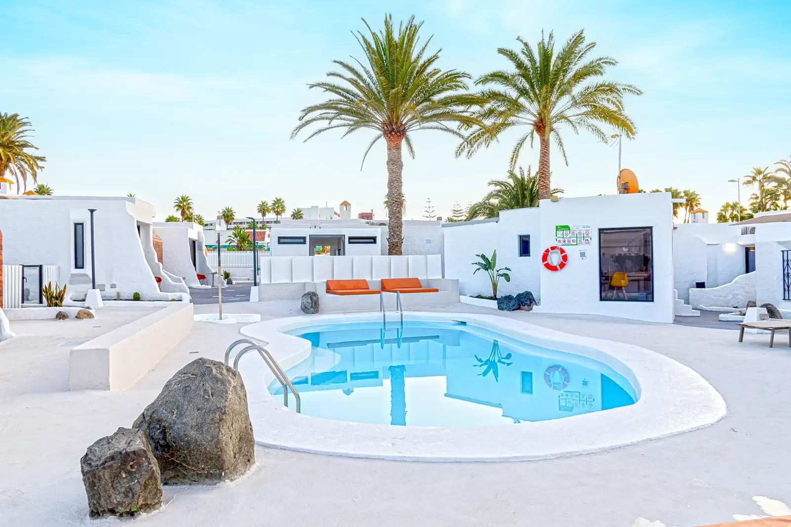 Een zwembad in een witte, mediterrane stijl resort met palmbomen, ligstoelen met oranje kussens en een helderblauwe hemel.