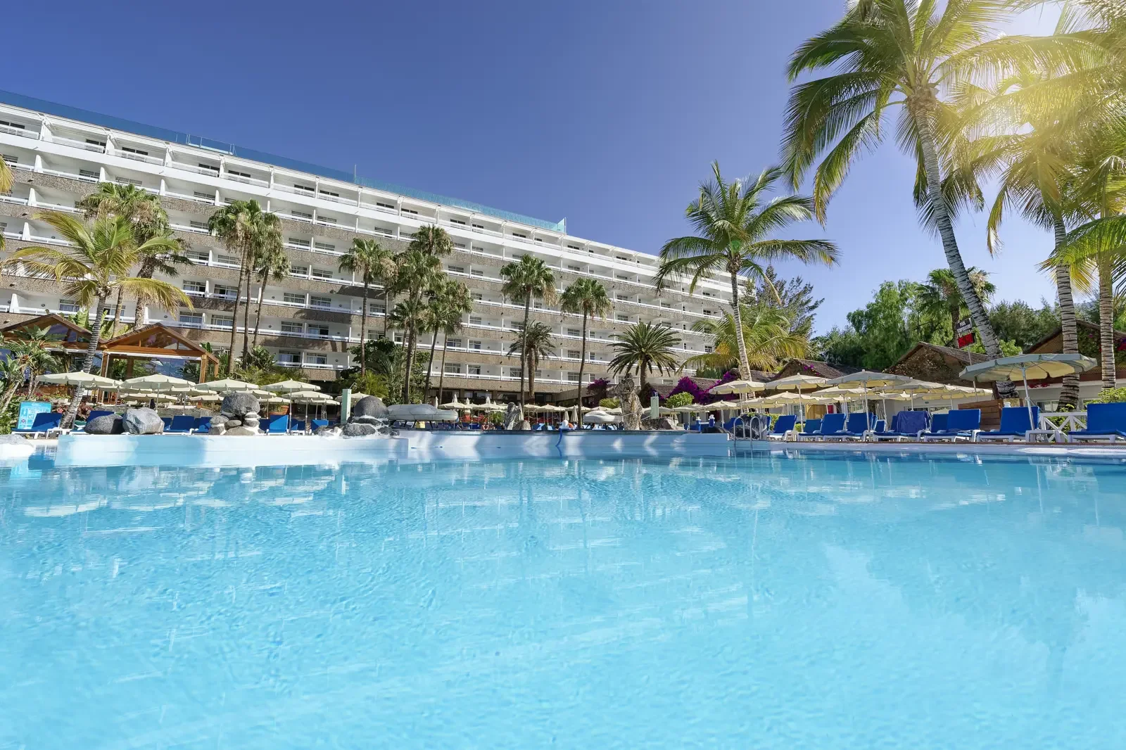 Zwembad met ligstoelen en parasols, palmbomen en een resort gebouw onder een heldere blauwe hemel.