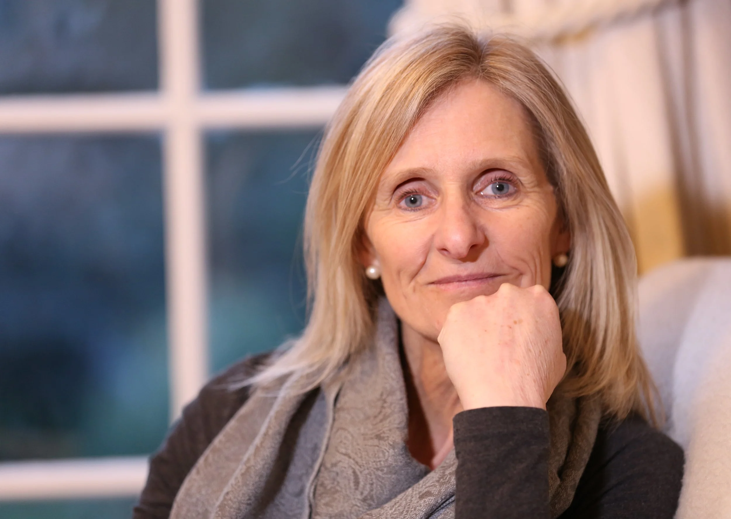 A woman with blonde hair and blue eyes resting her chin on her hand indoors.