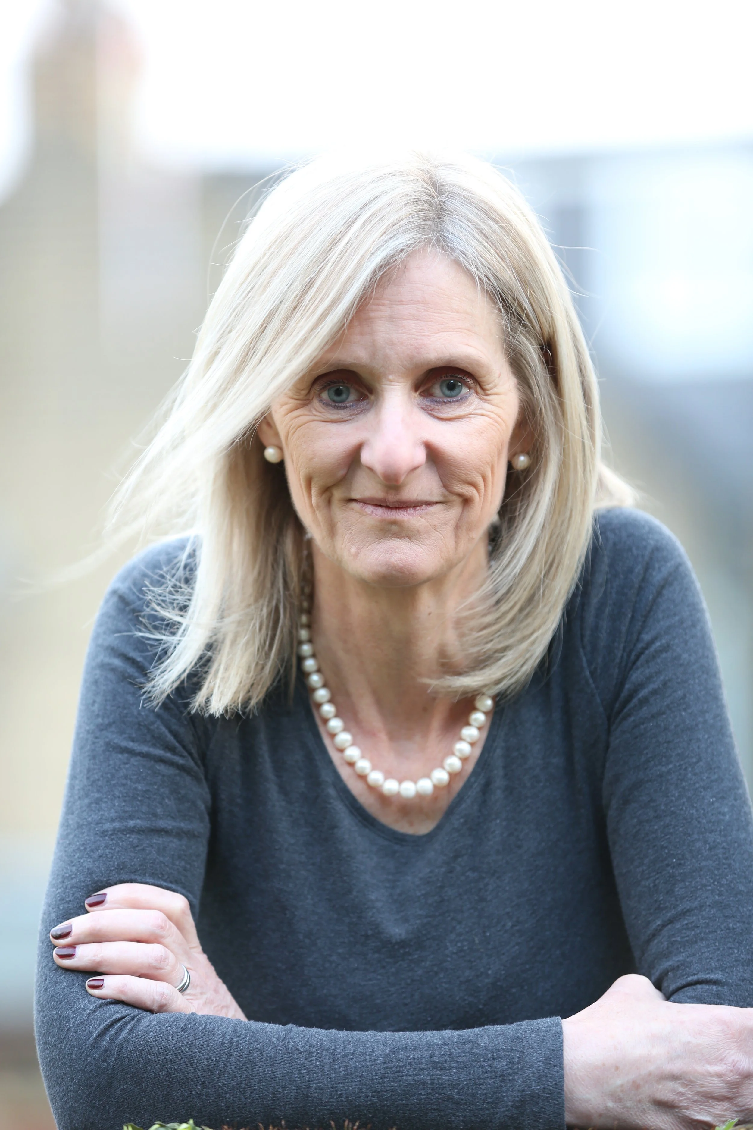 A middle-aged woman with blonde hair, blue eyes, wearing pearl earrings, a pearl necklace, and a dark gray long-sleeve shirt, is smiling and resting her arms on a surface outdoors.