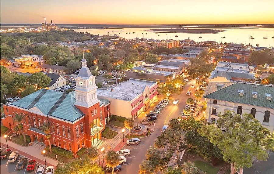 An aerial view of a downtown area at sunset, featuring a historic red brick building with a clock tower, surrounded by shops, trees, and parked cars, with a water body and ships in the background.