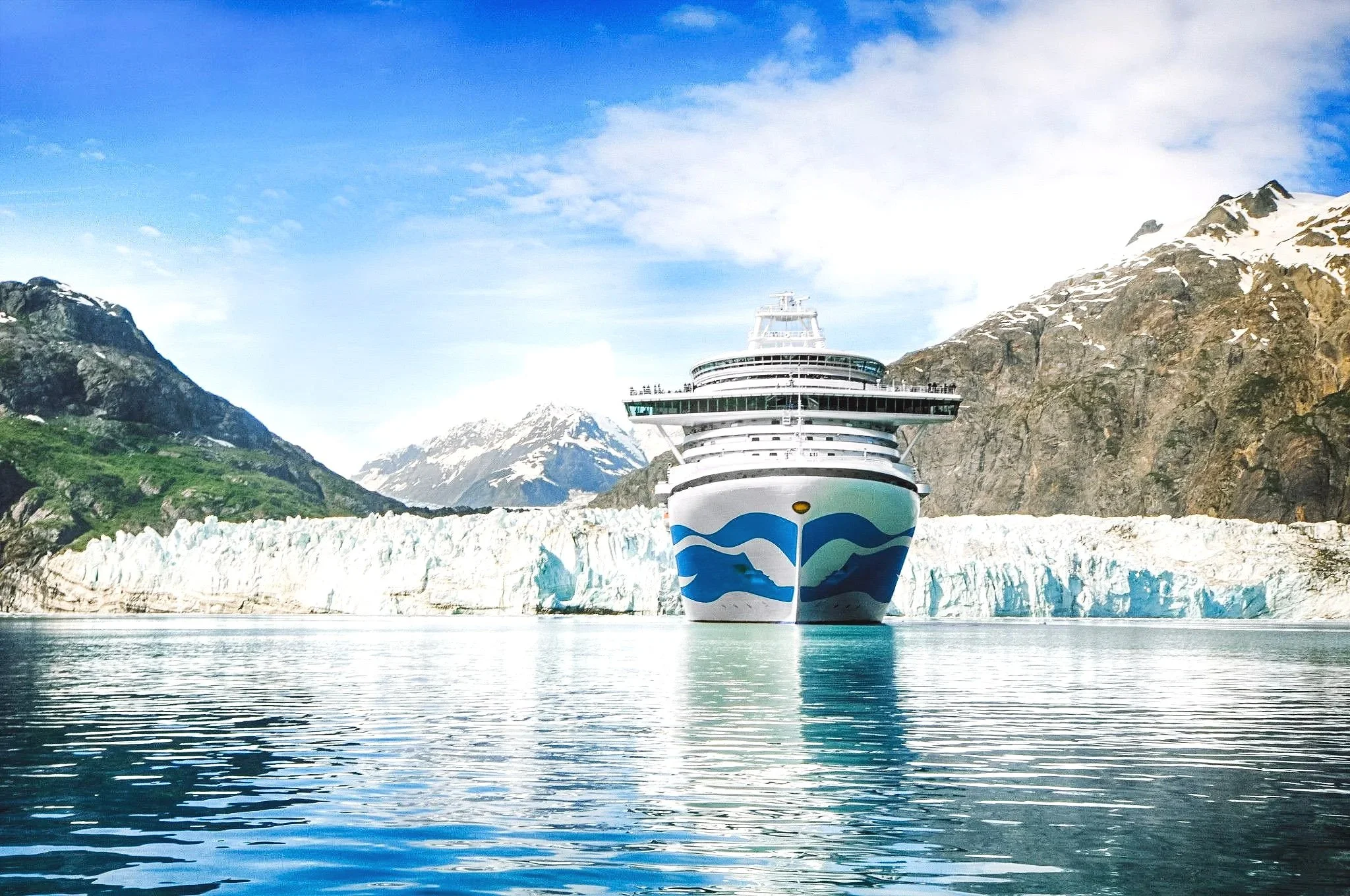 A cruise ship navigating through icy waters with snow-capped mountains in the background under a partly cloudy sky.