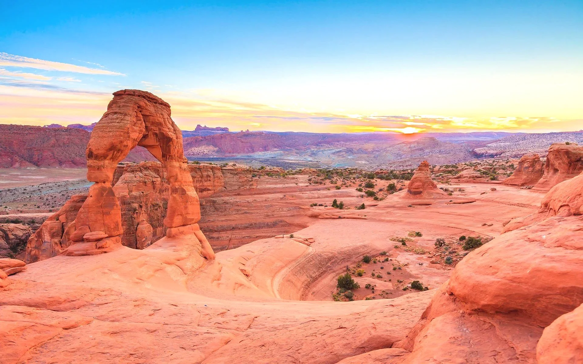 Sunset over red rock formations and the Delicate Arch in Arches National Park, Utah, with a colorful sky and distant mountains.
