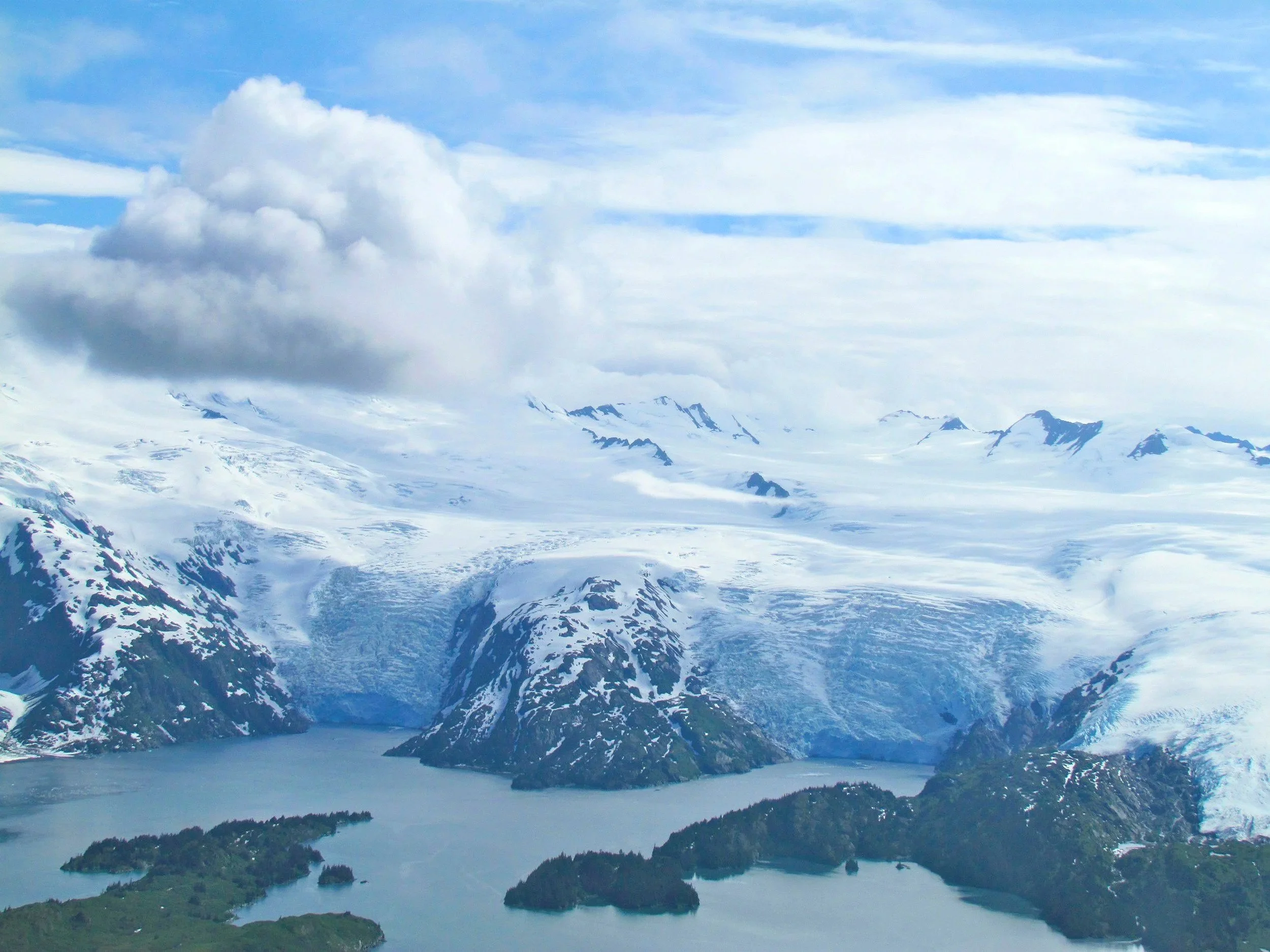 Snow-covered mountains and ice with clouds above a body of water and small forested islands in the foreground.