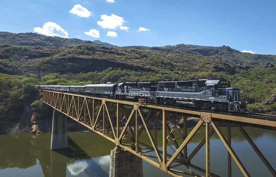 A train travels across a bridge over a river, with green hills and a partly cloudy sky in the background.