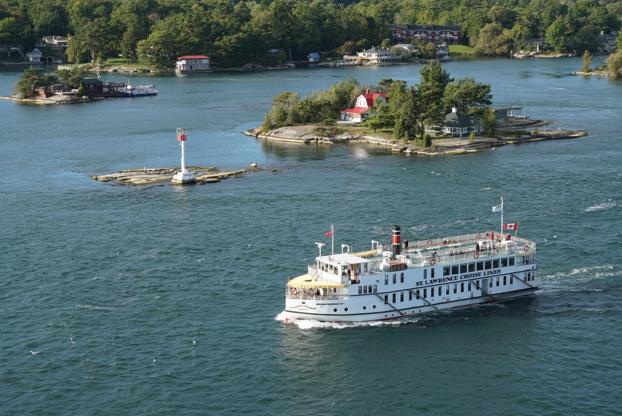 A white cruise boat labeled 'St. Lawrence Cruise Lines' sailing on a body of water near an island with trees and houses, with a small lighthouse on a rocky outcrop in the foreground.