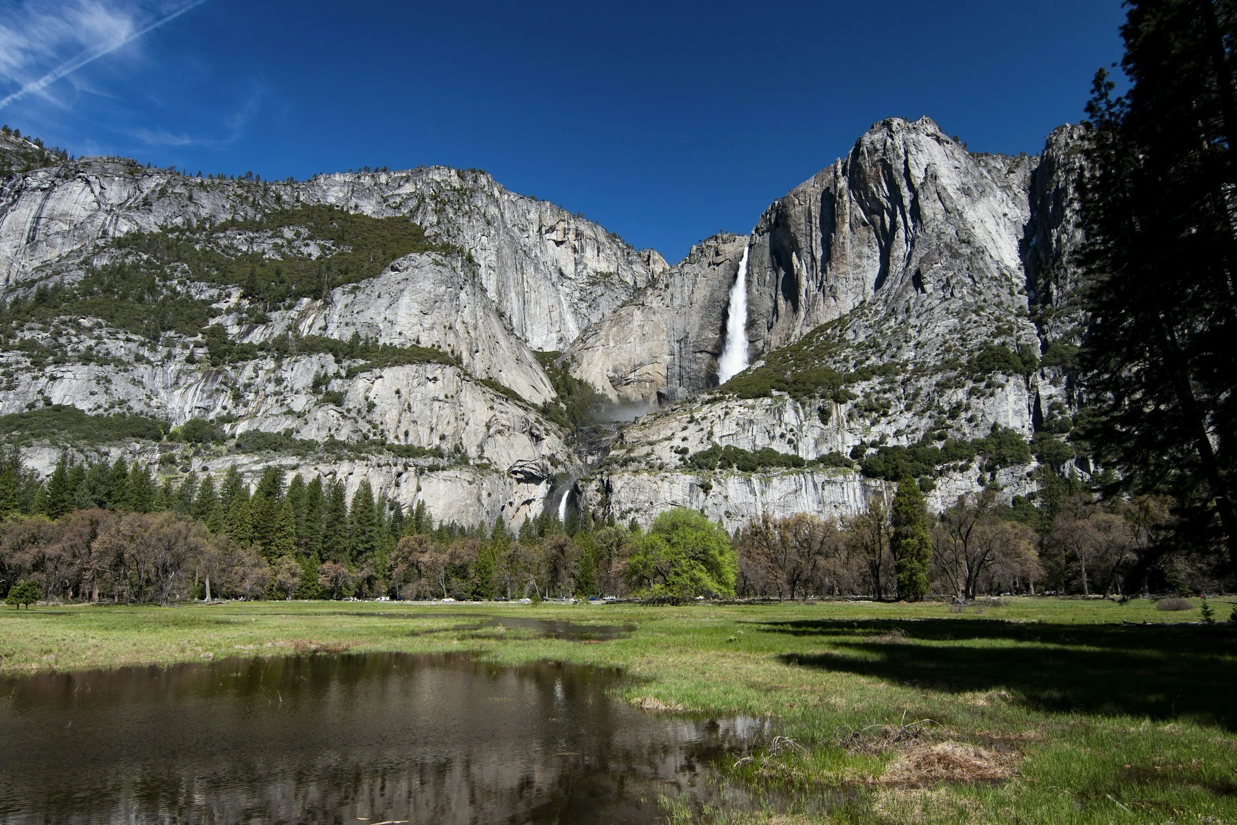 A scenic view of tall granite cliffs with a waterfall flowing down, surrounded by trees and a grassy field with a pond in the foreground under a clear blue sky.
