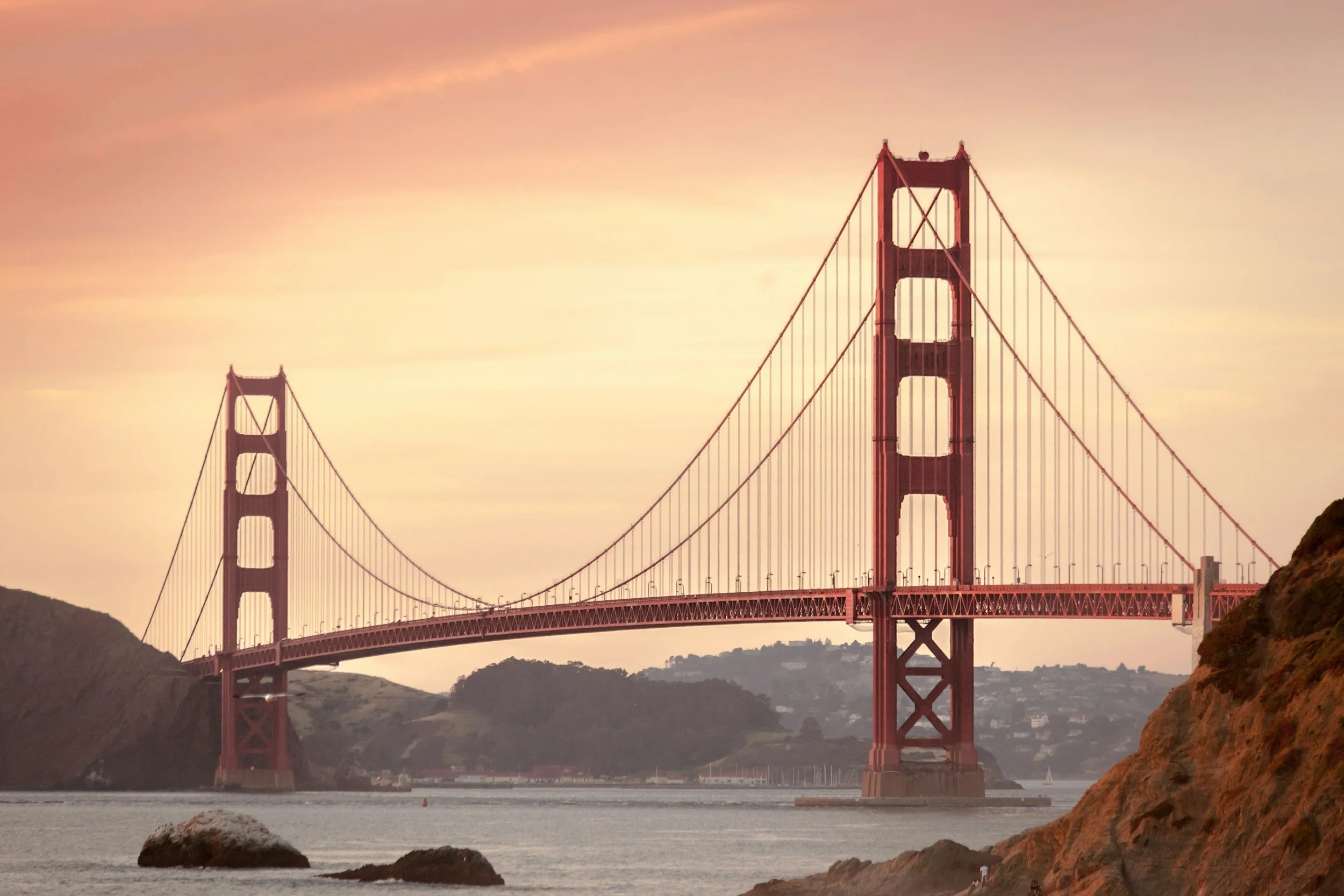 View of the Golden Gate Bridge in San Francisco at sunset, with the sky in pink and orange hues, and water in the foreground with rocks along the shoreline.