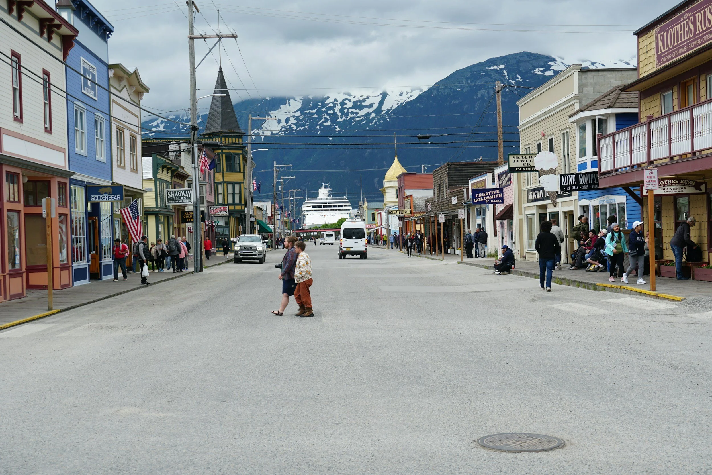 Main street in a small town with colorful buildings, people walking and shopping, a cruise ship in the distance, mountains with snow, and cloudy sky.