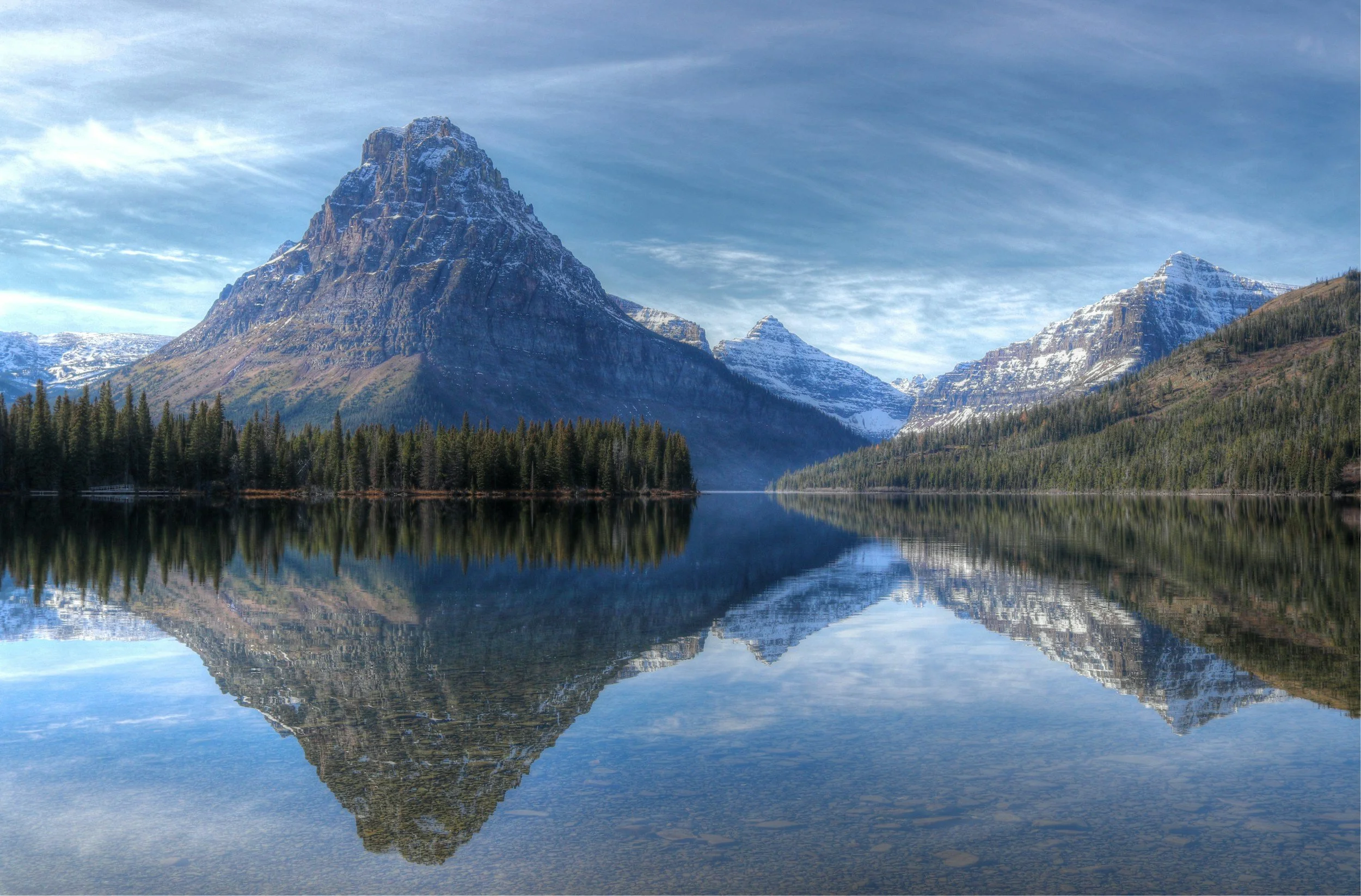 Snow-capped mountains reflected in a calm lake with a forest at the shoreline under a blue sky with wispy clouds.