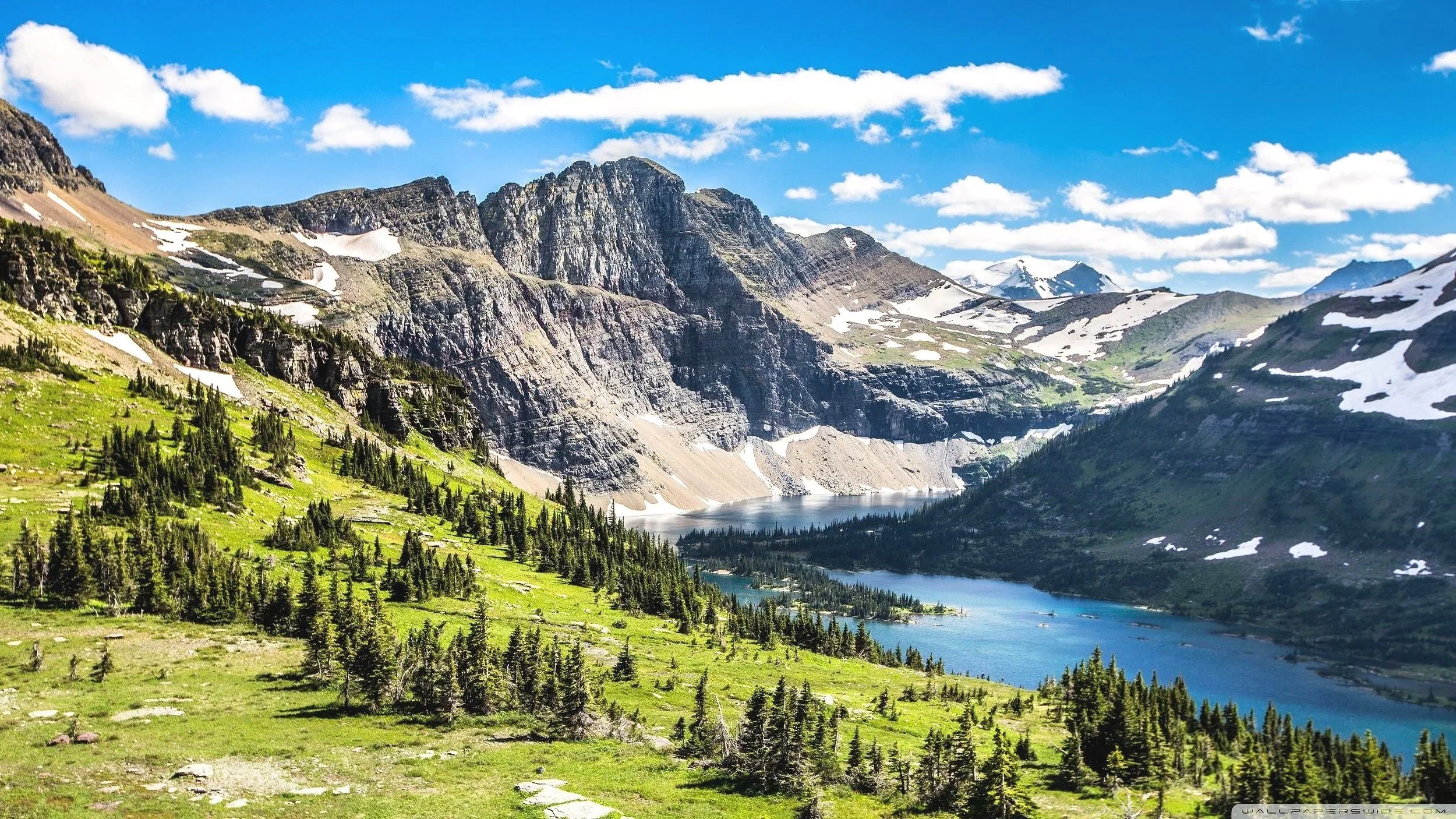 A scenic mountain landscape with a lake in the valley, surrounded by lush green trees and rugged snow-capped mountains under a partly cloudy blue sky.