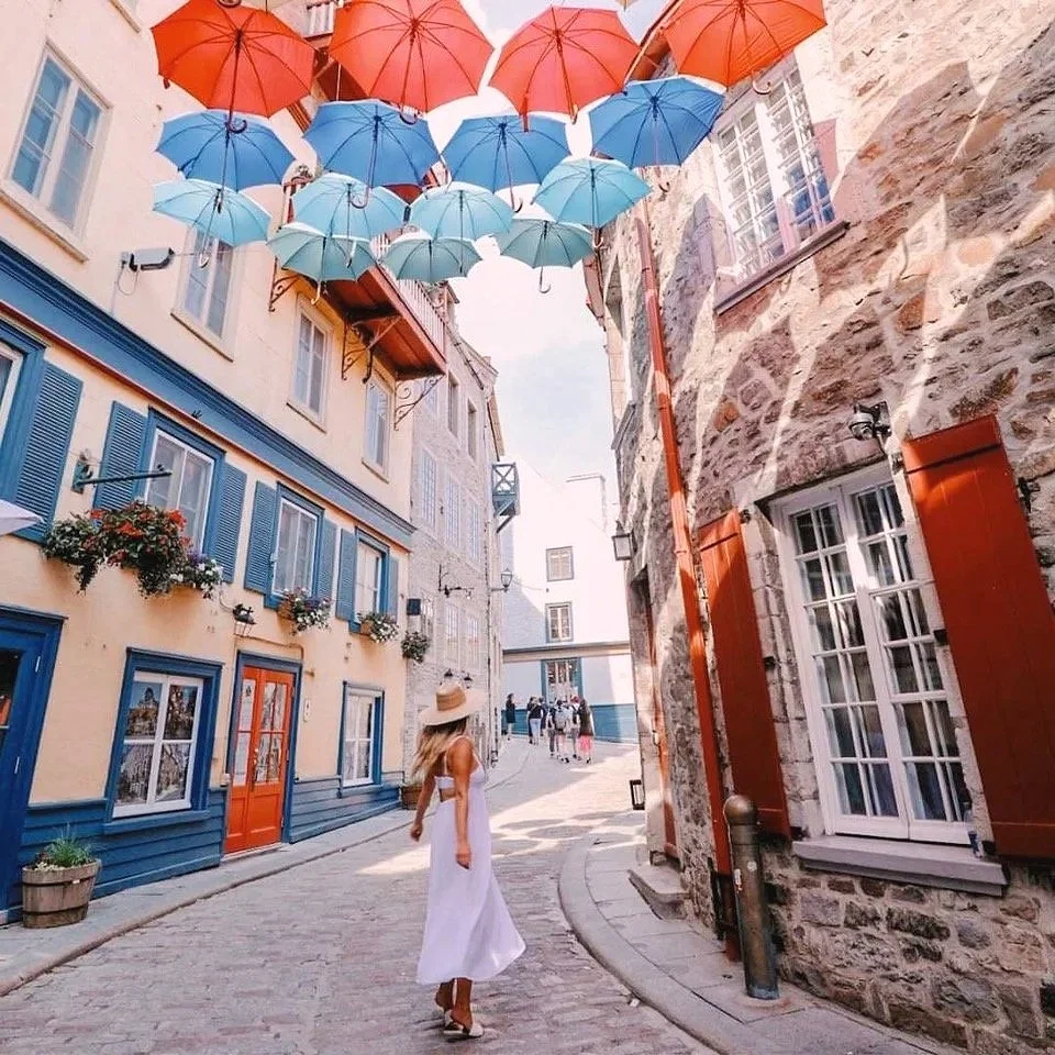 Colorful umbrellas hanging overhead in a narrow street with stone and pastel-colored buildings. A woman dressed in white with a wide-brimmed hat is walking on the cobblestone street.