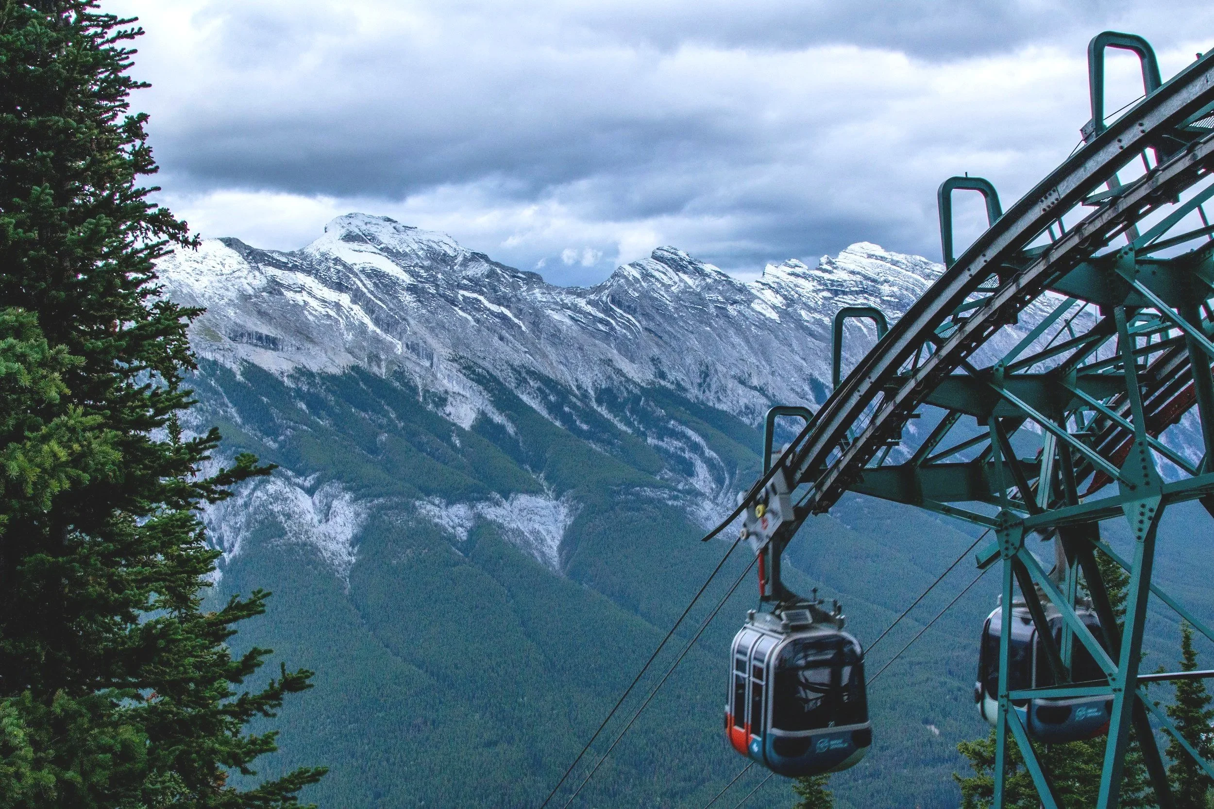 A scenic view of snow-capped mountains under a cloudy sky with a section of a roller coaster track and a gondola lift in the foreground, and a green pine tree on the left.