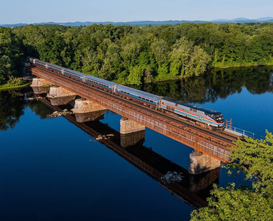 A modern passenger train traveling across a metal bridge over a calm river with green trees on both sides and mountains in the background.