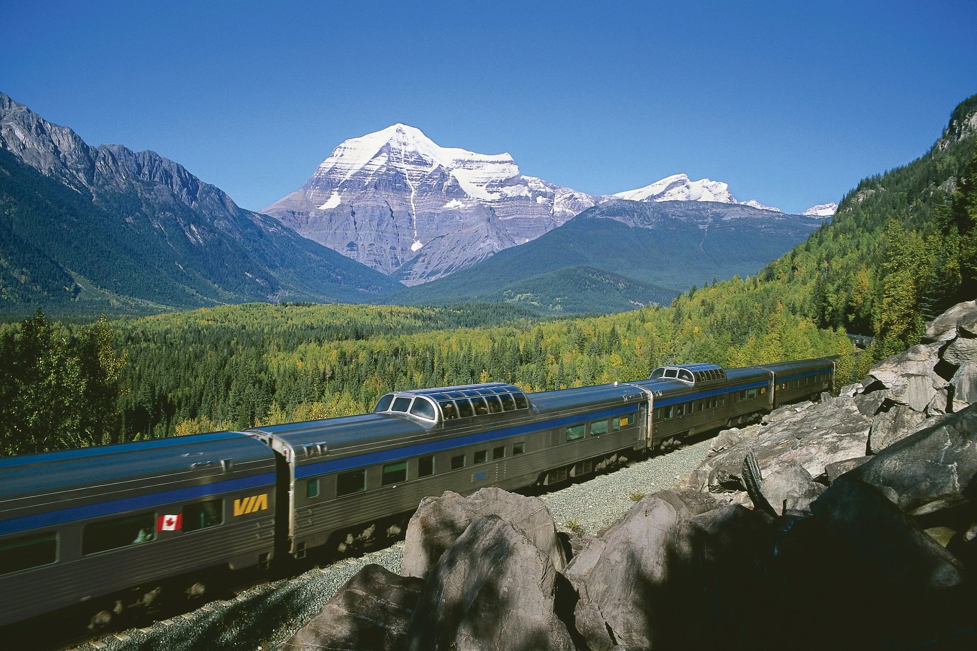 Snow-capped mountains and a green forest with a train traveling along the tracks.