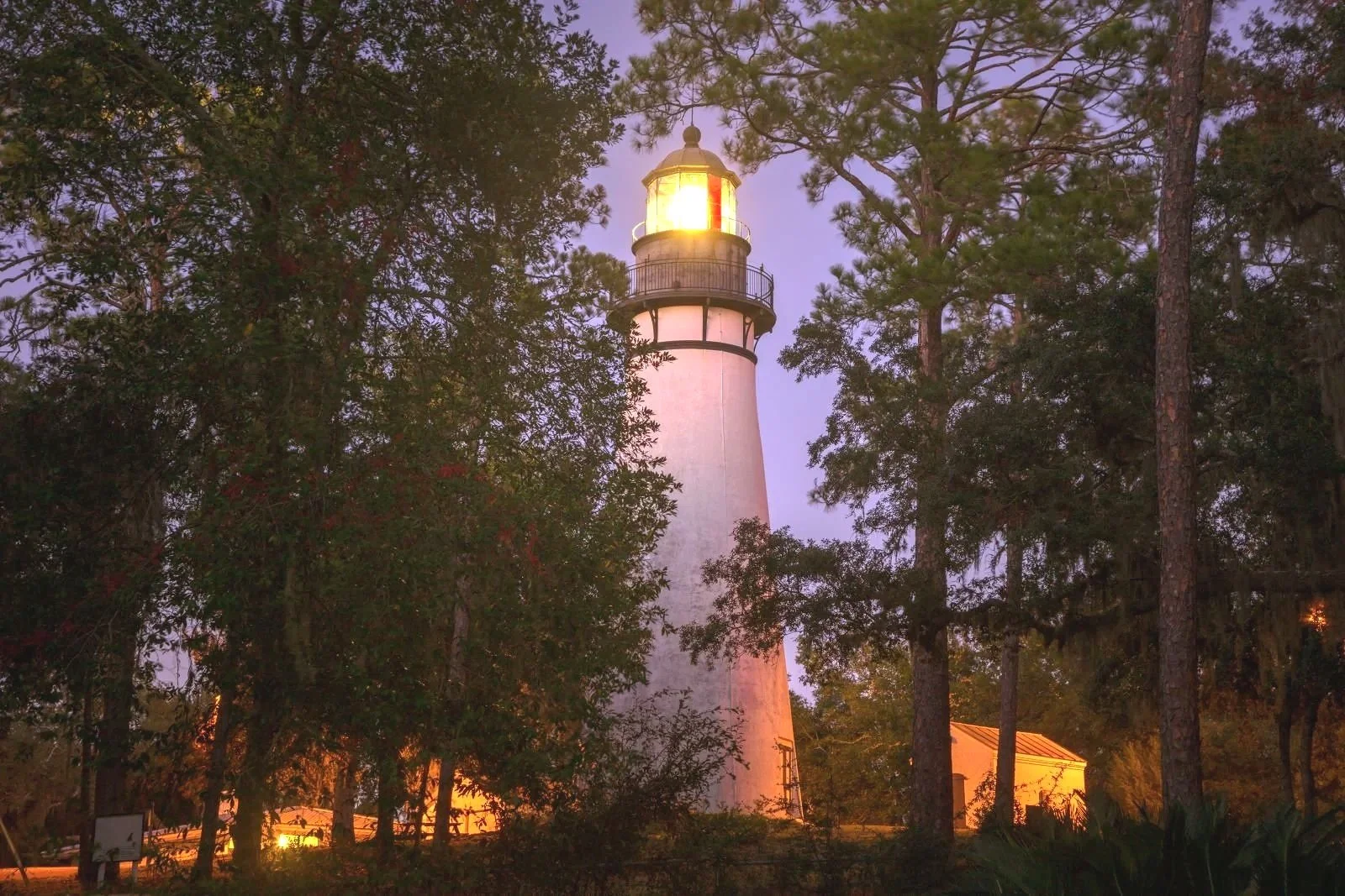 A lighthouse emitting light through its top window, surrounded by trees at dusk or dawn.
