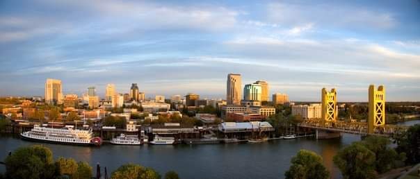 Panoramic view of a city skyline with tall buildings, a river, and a bridge with two prominent yellow towers, under a partly cloudy sky during daytime.