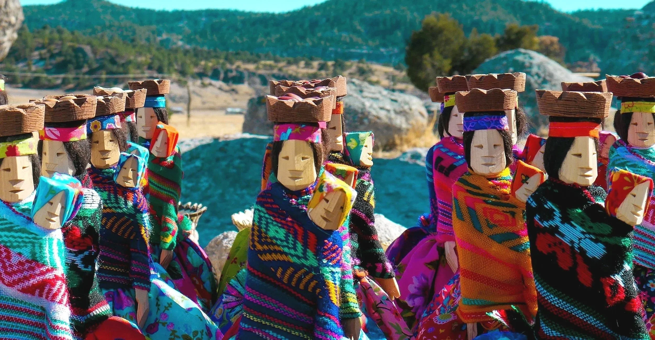 Colorful traditional dolls with wooden faces, wearing embroidered clothing and hats, standing outdoors against a mountainous landscape.