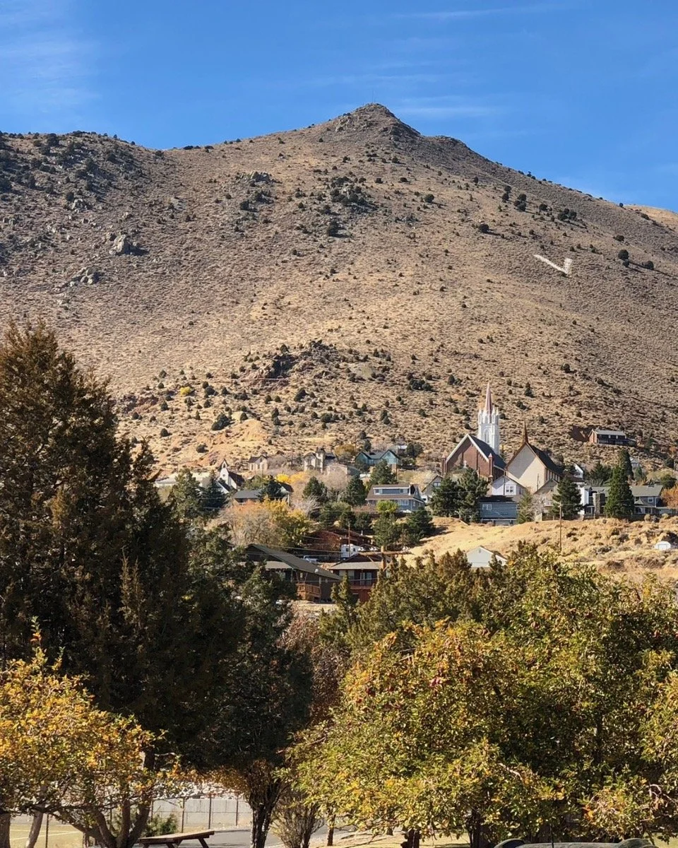 A small town with houses and a church with a tall steeple at the base of a rocky, brown mountain under a blue sky.