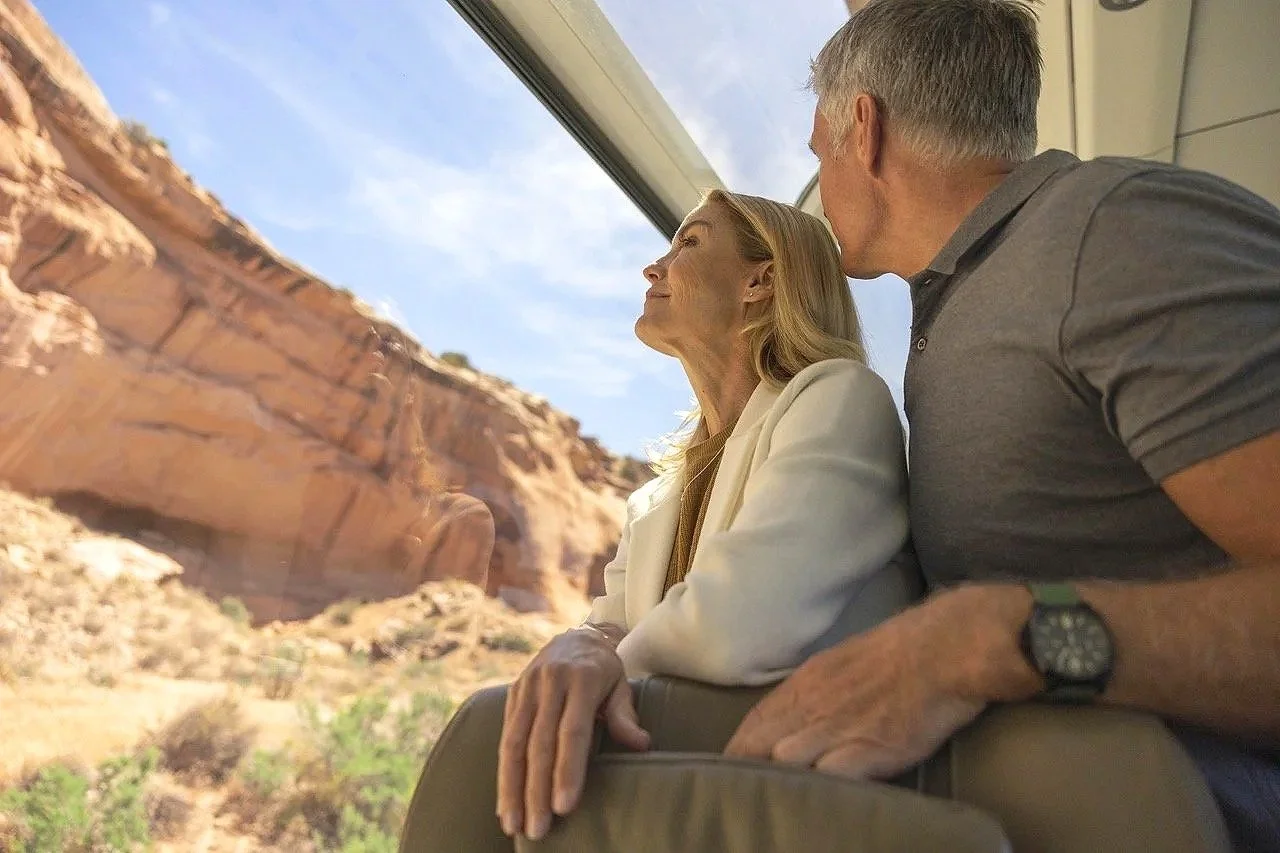 An elderly woman and man looking out of a large window at a desert landscape with rocky cliffs.