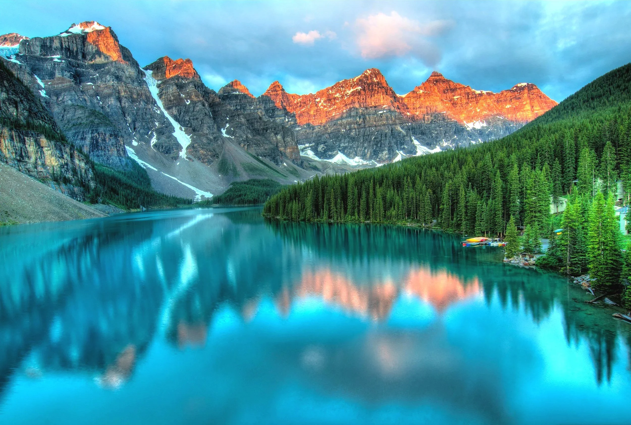 A scenic mountain landscape with snow-capped peaks illuminated by the setting sun, a calm turquoise lake reflecting the mountains, a dense forest of pine trees, and colorful kayaks on the lake shore.