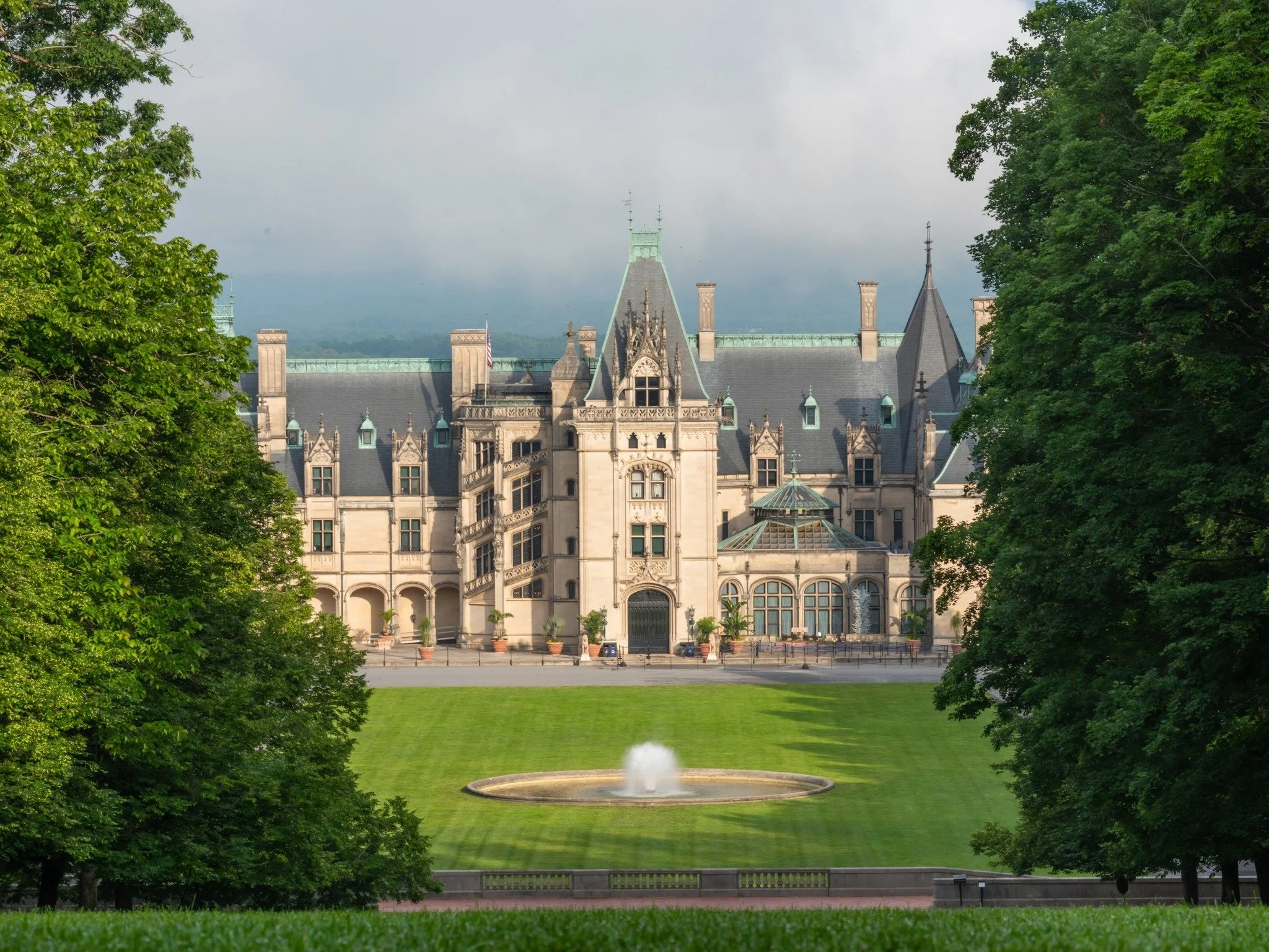 View of a historic castle-like building with pointed roofs and ornate architecture, framed by lush green trees and a fountain in the foreground.
