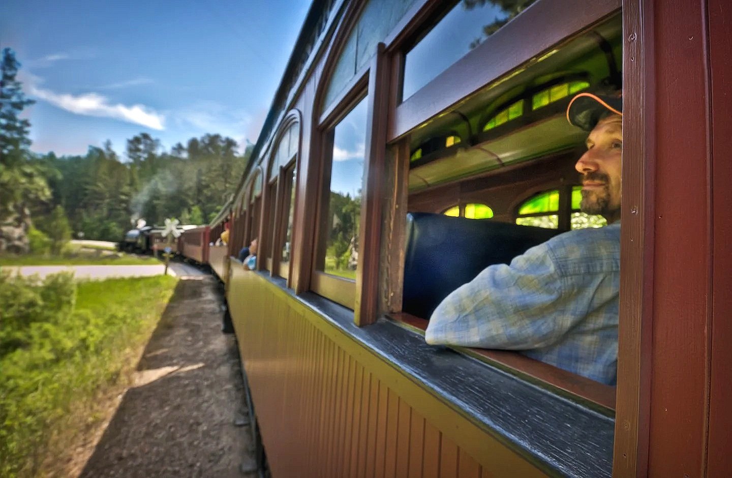 A man wearing a baseball cap and plaid shirt looking out the window of a vintage train car as the train travels through a lush green landscape on a sunny day.
