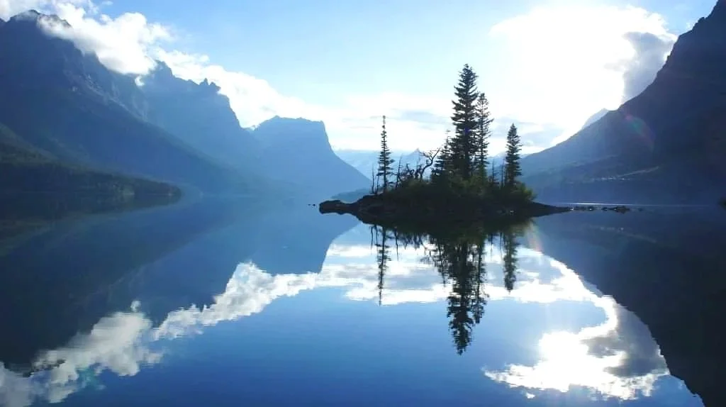 A small island with trees in a calm mountain lake, reflecting the surrounding mountains and sky.