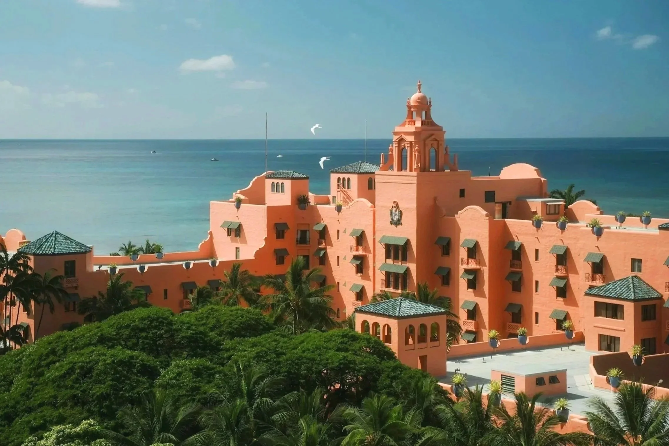 A pink-colored building with towers and terraces overlooking the ocean, surrounded by green trees and with boats on the water in the background under a partly cloudy sky.