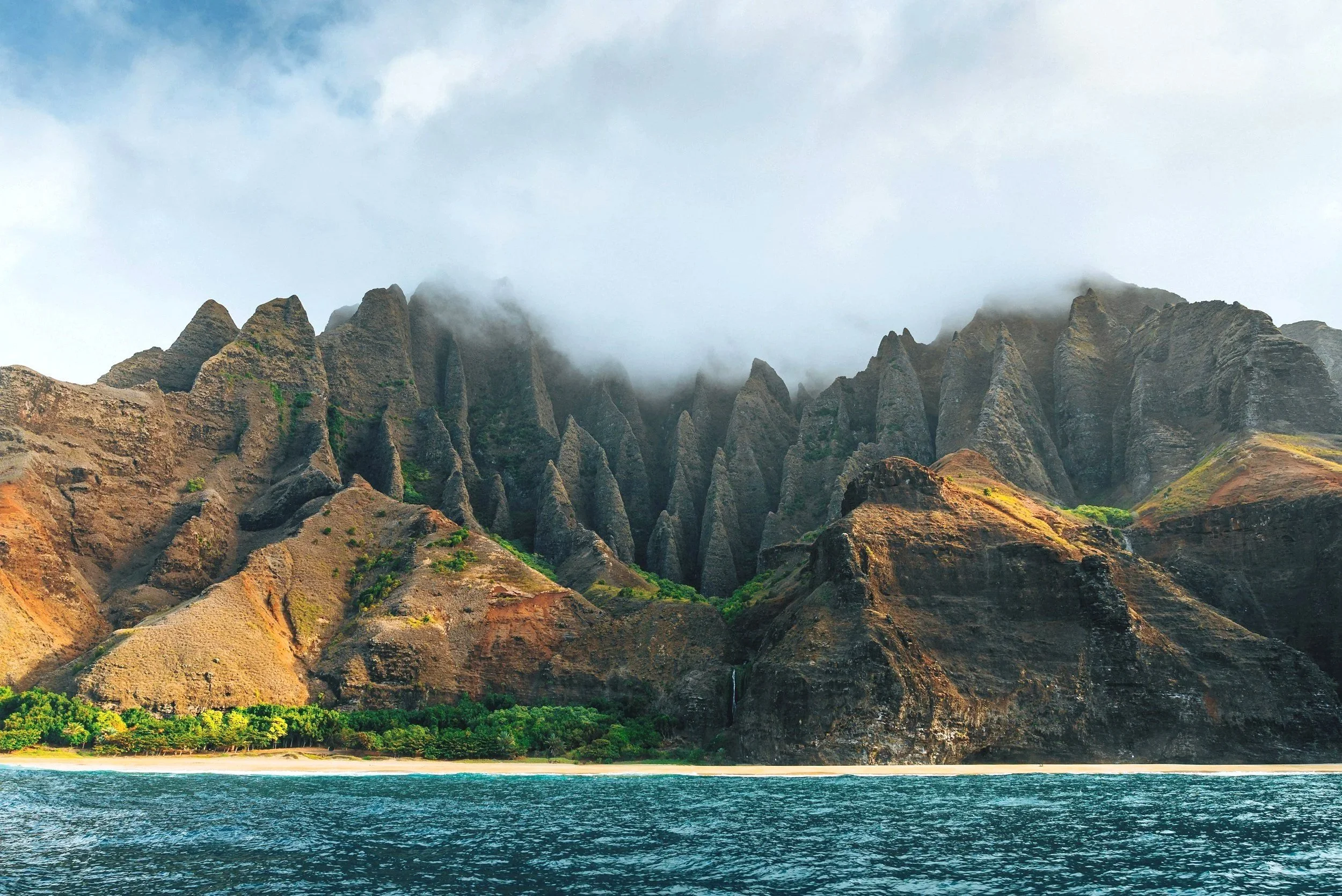 A scenic view of rugged mountains with mist covering their peaks, a lush green forest below, and a body of water in the foreground.