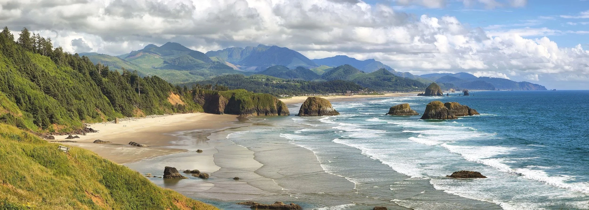 Beach with sandy shoreline, large rocks in the water, green forested hills, and mountains in the background under cloudy sky.