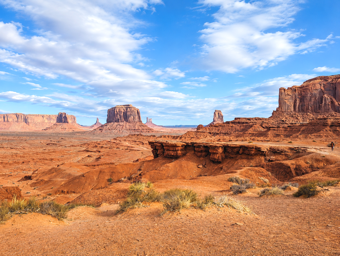 Desert landscape with large red rock formations, scattered vegetation, and a partly cloudy blue sky.