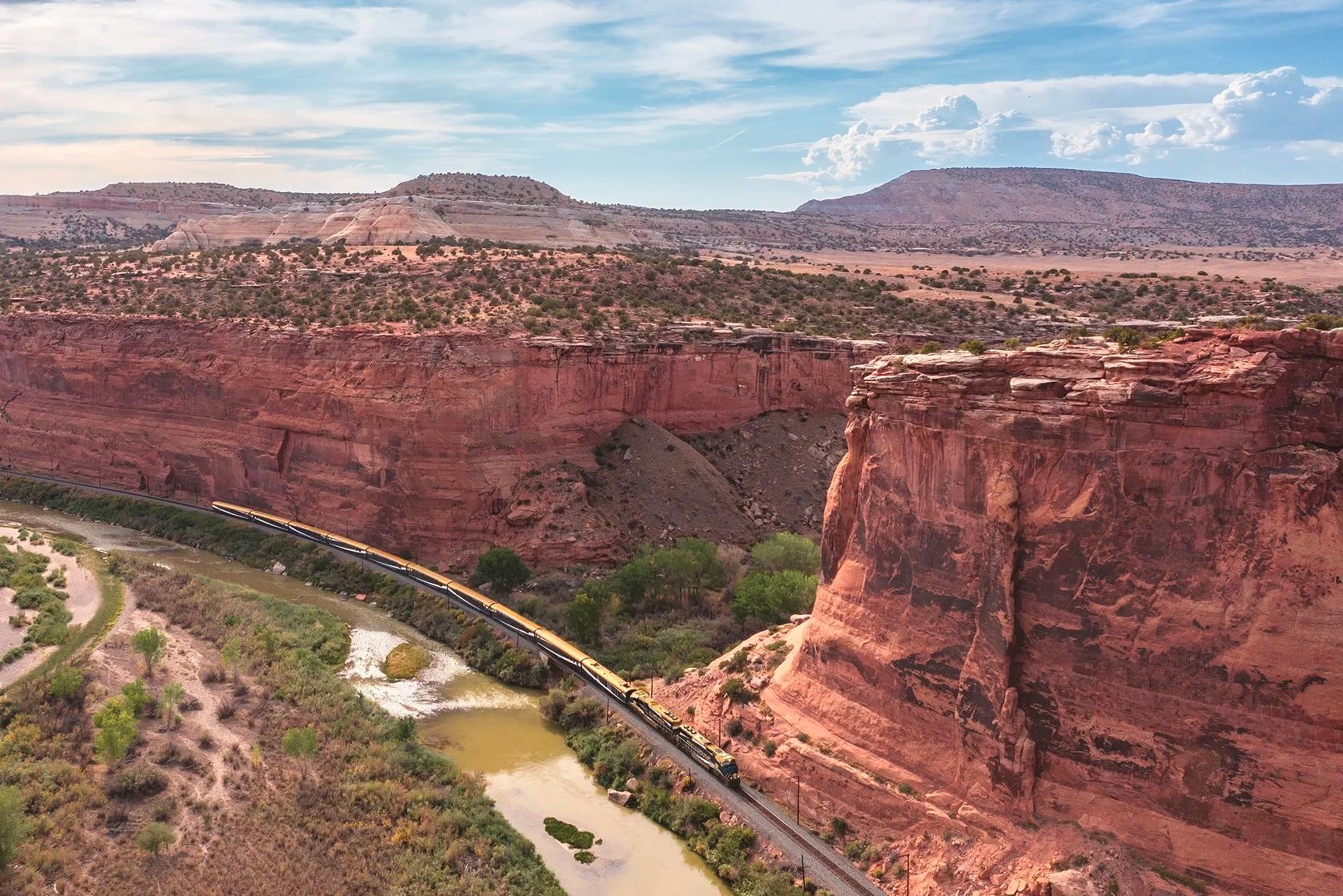 A train traveling through a canyon with red rock cliffs and a river at the bottom, under a blue sky with clouds.