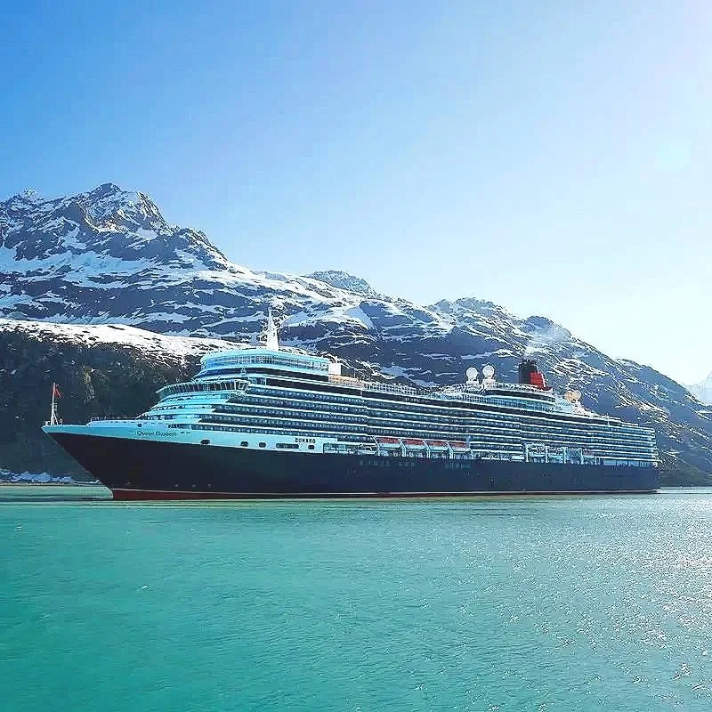 Large cruise ship docked near snow-covered mountains with clear blue sky and calm water.