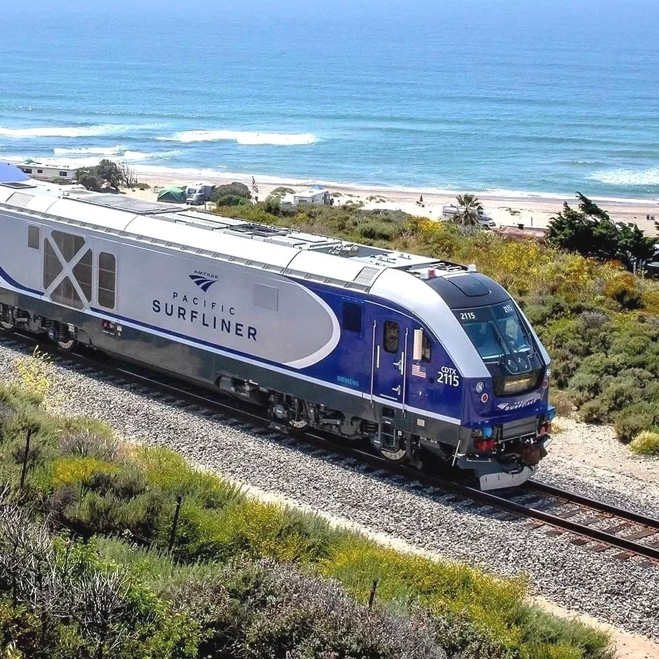 A Pacific Surfliner train traveling along a coastal track with beach and ocean in the background.