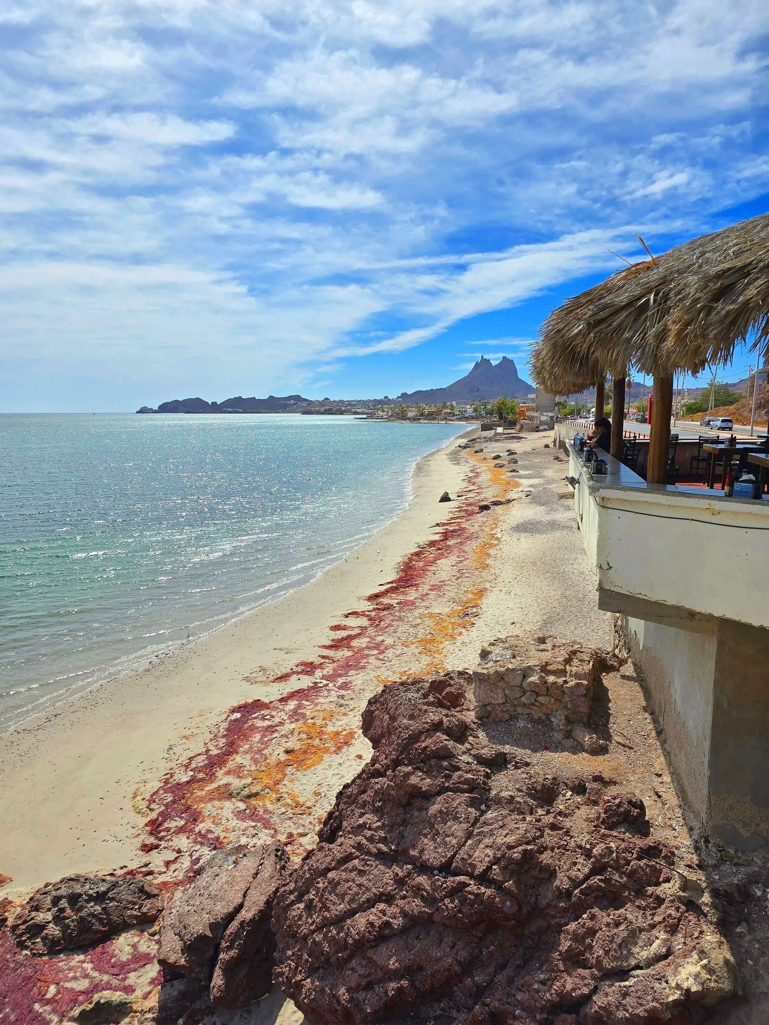 A beach with rocks, a thatched umbrella, and a view of distant mountains against a blue sky with clouds.