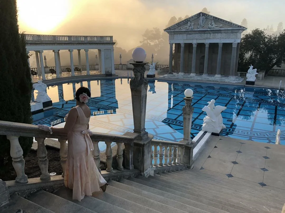 A woman in a light-colored dress and with a flower in her hair standing on stairs near a decorative pool at sunrise or sunset with classical-style sculptures and structures in the background.