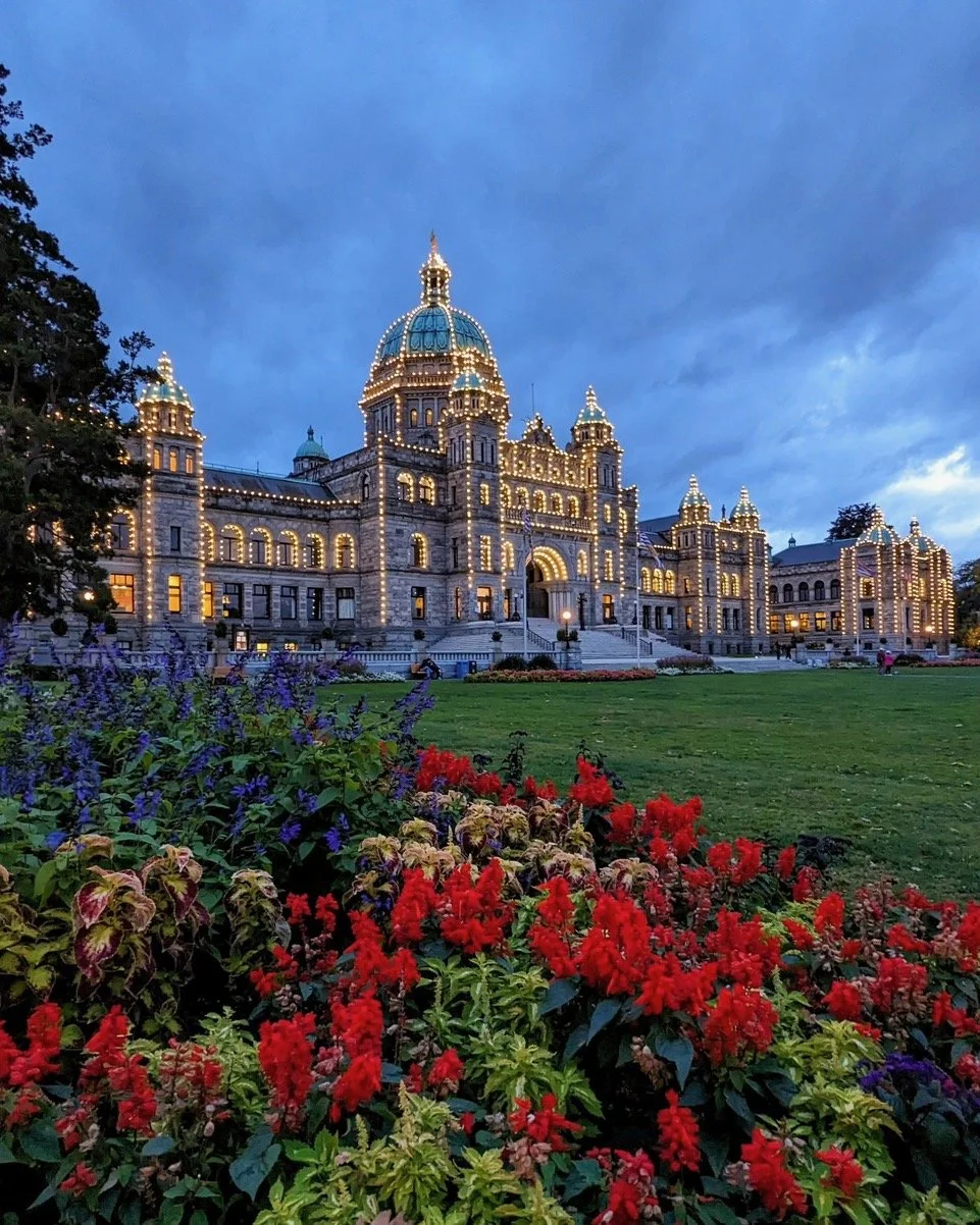 View of the Idaho State Capitol building illuminated at dusk, with colorful flower beds in the foreground and a cloudy sky overhead.