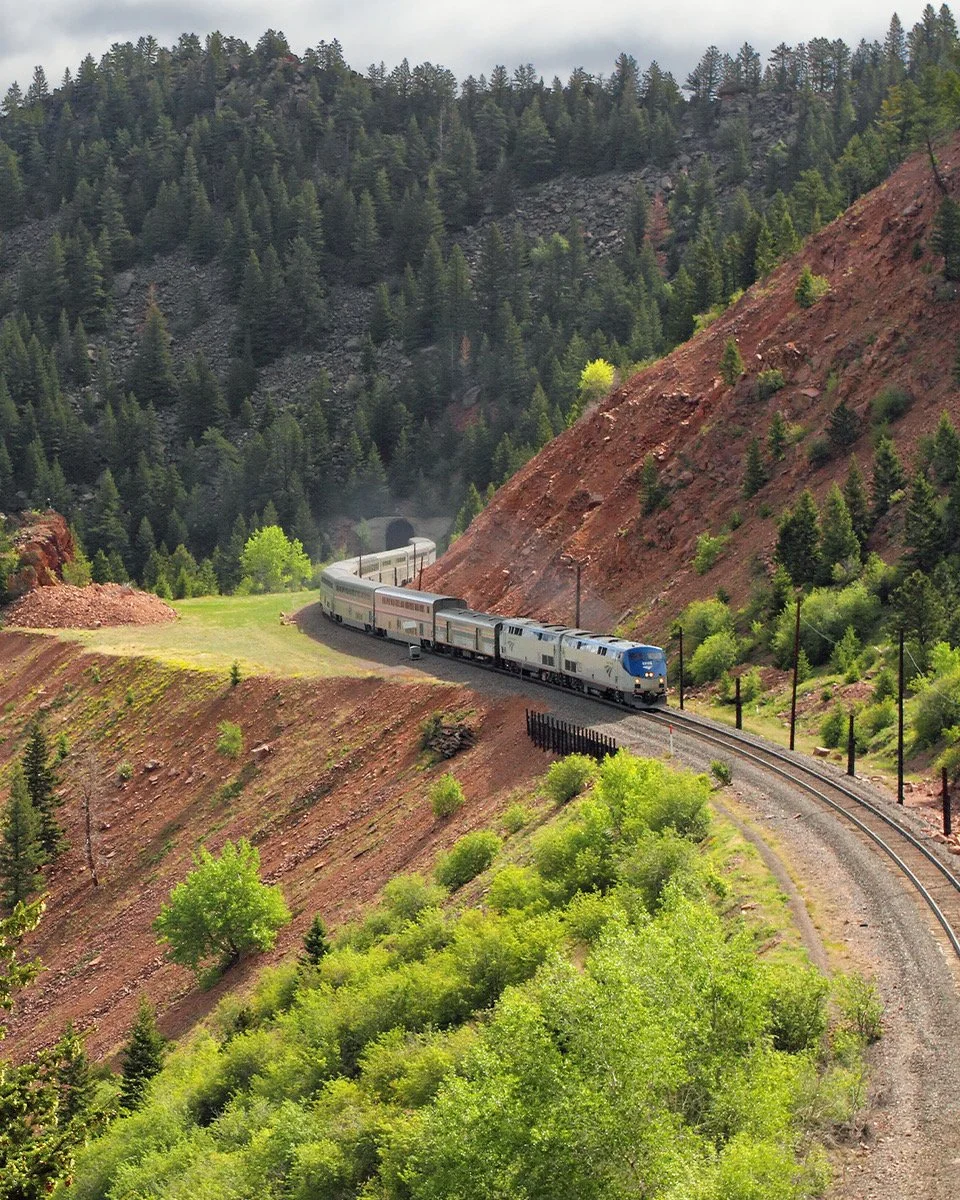 A scenic mountain landscape with a train traveling through a green valley, surrounded by dense forest and rocky hills.