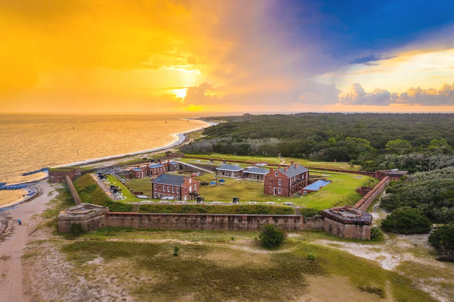 Aerial view of historic brick fort with cannons, green grass, and pathways, overlooking a beach and ocean at sunset, with a cloudy sky and distant shoreline.