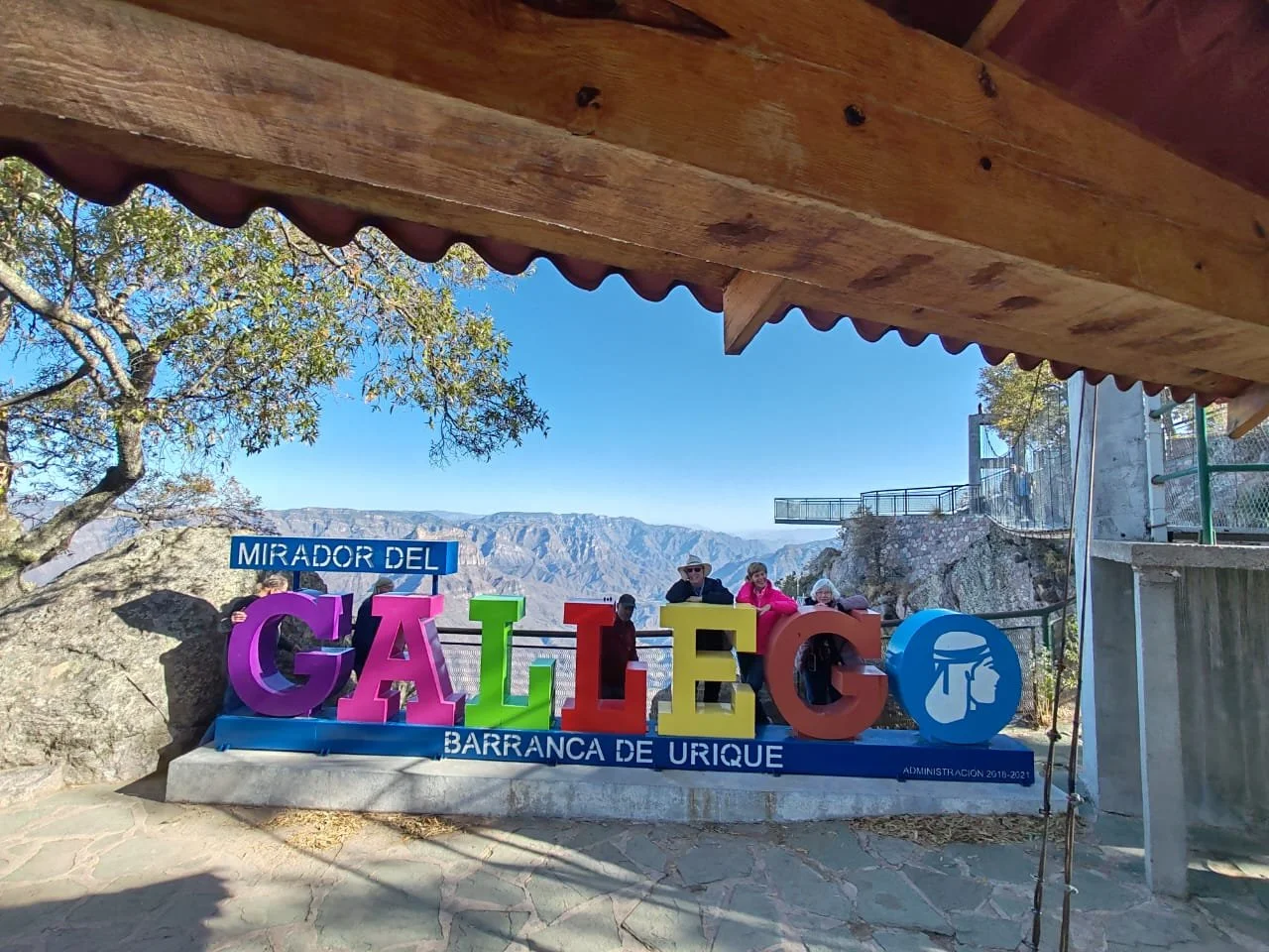 Colorful sign reading 'GALLITO' with smaller signs 'MIRADOR DEL' above and 'BARRANCA DE URIQUE' below, set outdoors with mountains in the background, under a wooden roof.