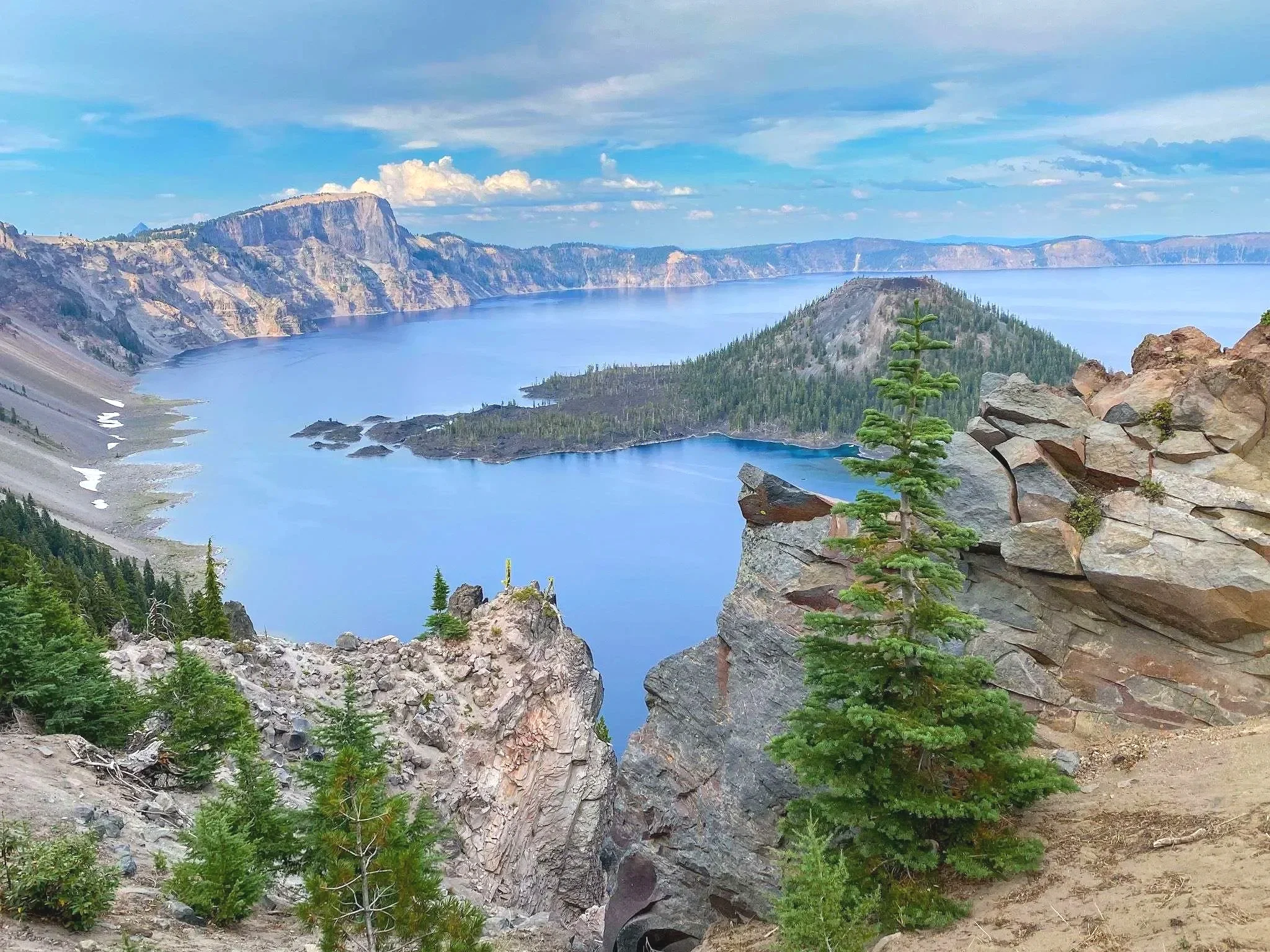 View of a large blue lake surrounded by steep rocky cliffs and green pine trees, with cloudy sky above.